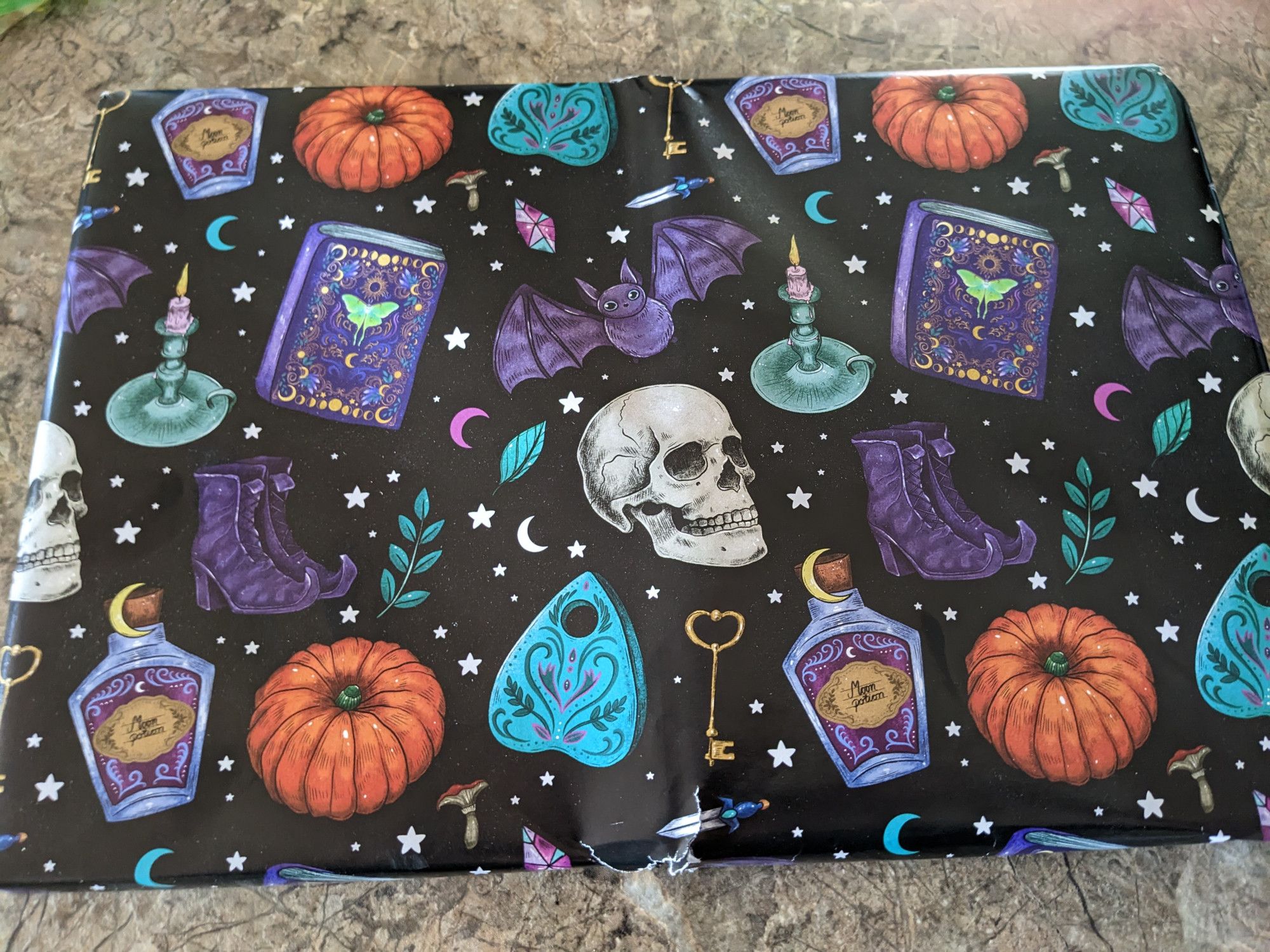 A rectangular package. The wrapping paper is black with colourful pumpkins, ouija board planchettes, bats, boots, skulls, candles, leaf sprigs, small crescent moons, bottles of poison, and grimoires featuring luna moths.