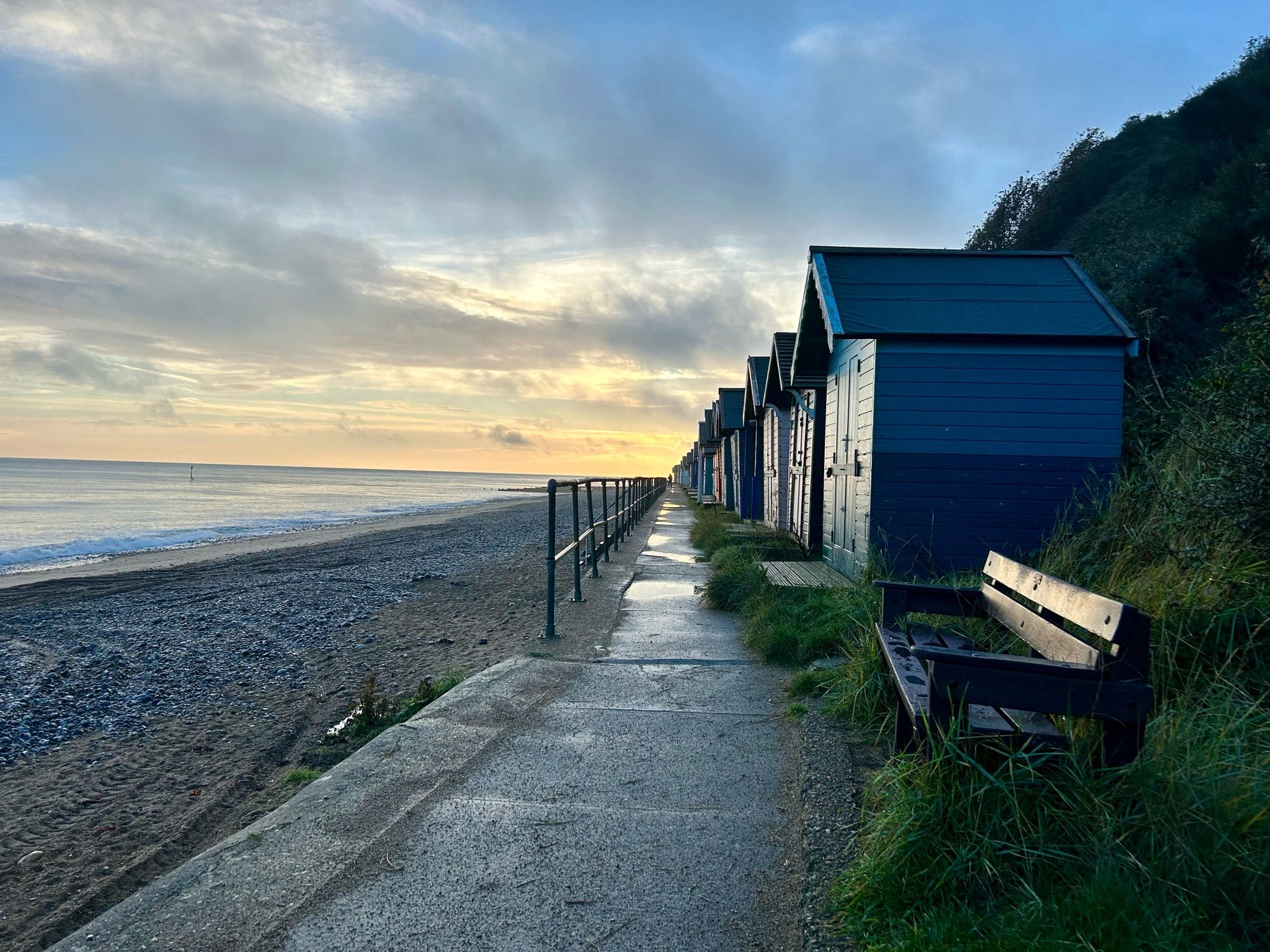 A waiting bench and tumbled row of beach huts face the sea and catch the morning light. An equally tumbled sky’s light catches last night’s rain. The sea gives back the sky.