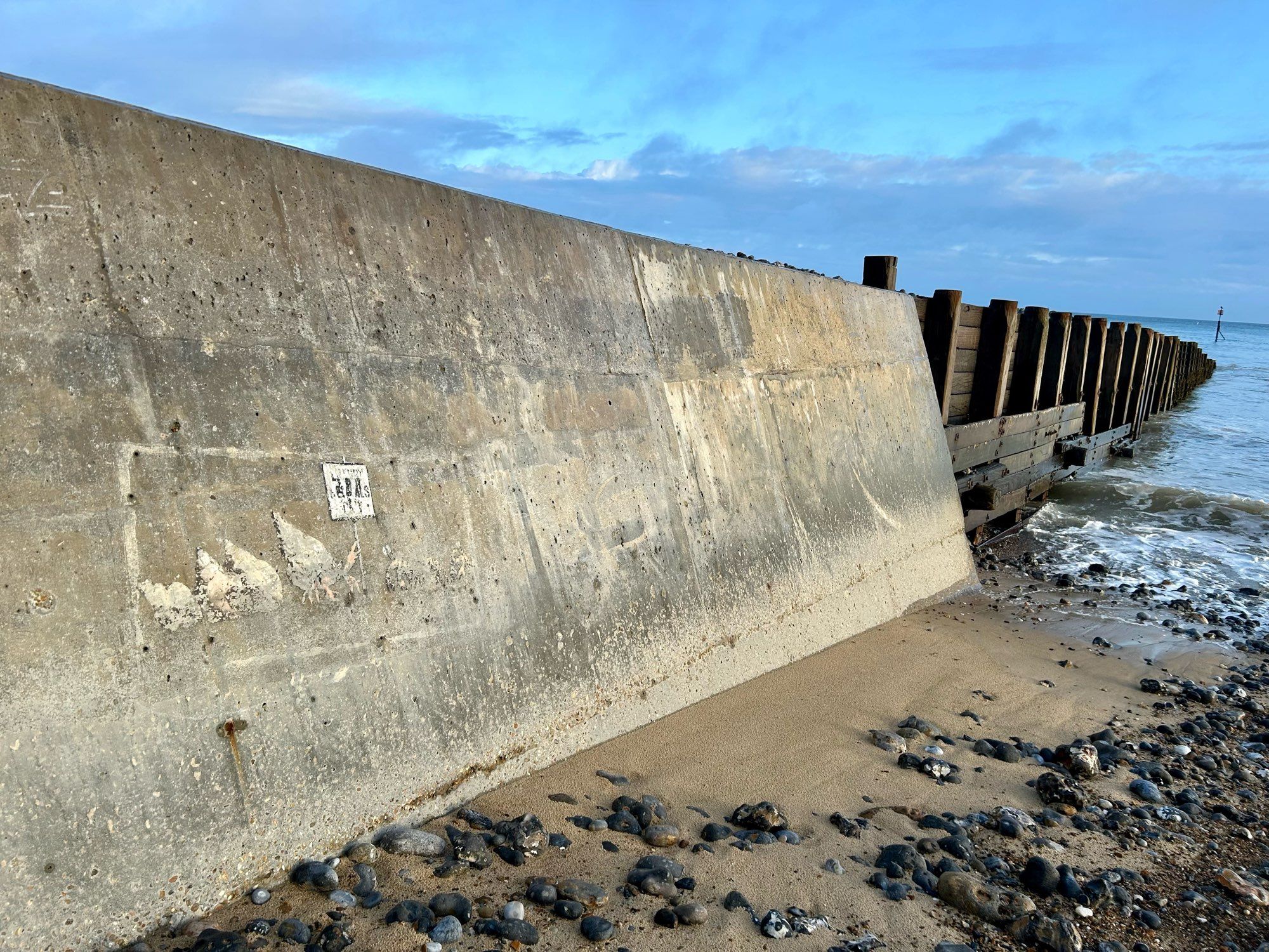 A weathered Banksy from 2021, faded hermit crabs hold a sign you can no longer read. On a concrete groyne which becomes wooden as it reaches the waves and disappears. https://banksyexplained.com/a-great-british-spraycation-august-2021/