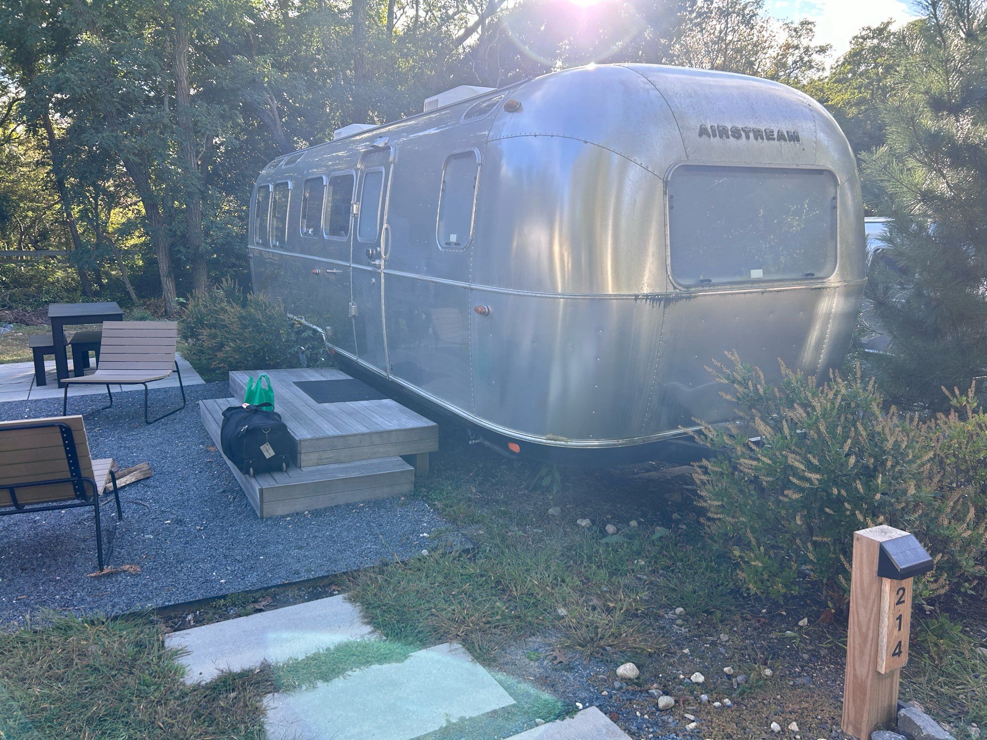 Photo of my home last week - a silver “Airstream” camper in a forested area, beside an outdoor firepit. Seating and a table are near the fire, and wooden steps have been built under the Airstream door.