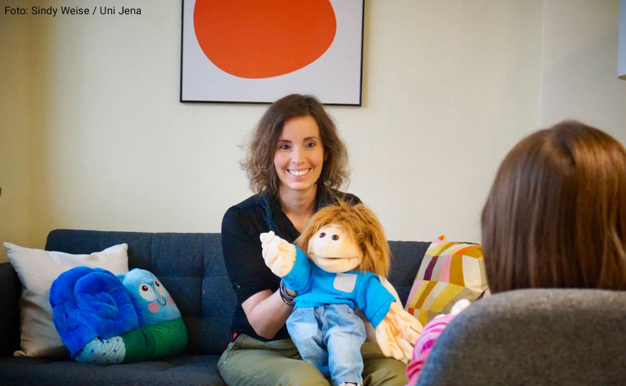 Prof. Dr Julia Asbrand from the University of Jena sits on a sofa opposite a child and uses a doll during therapy.