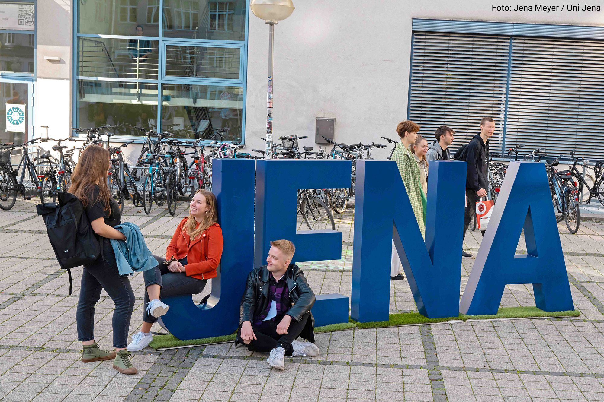 Große blaue Buchstaben bilden auf dem Campus Ernst-Abbe-Platz das Wort Jena. Eine Gruppe Studierender läuft über den Campus, drei andere Studierende unterhalten sich.