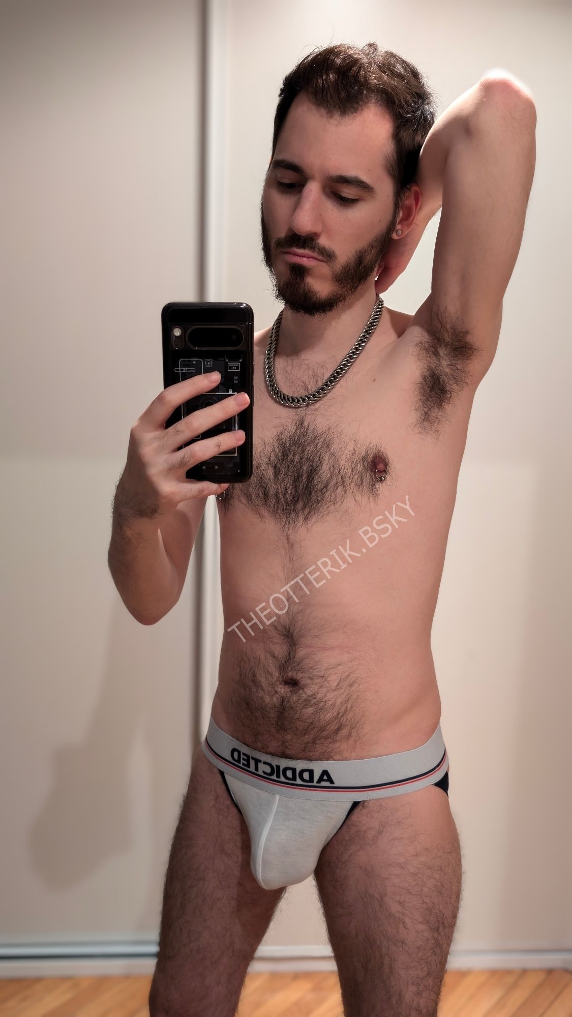 Mirror selfie of a white man in his early 30s just wearing a white jockstrap and a chain necklace. He is hairy and has a beard and nipple piercings.