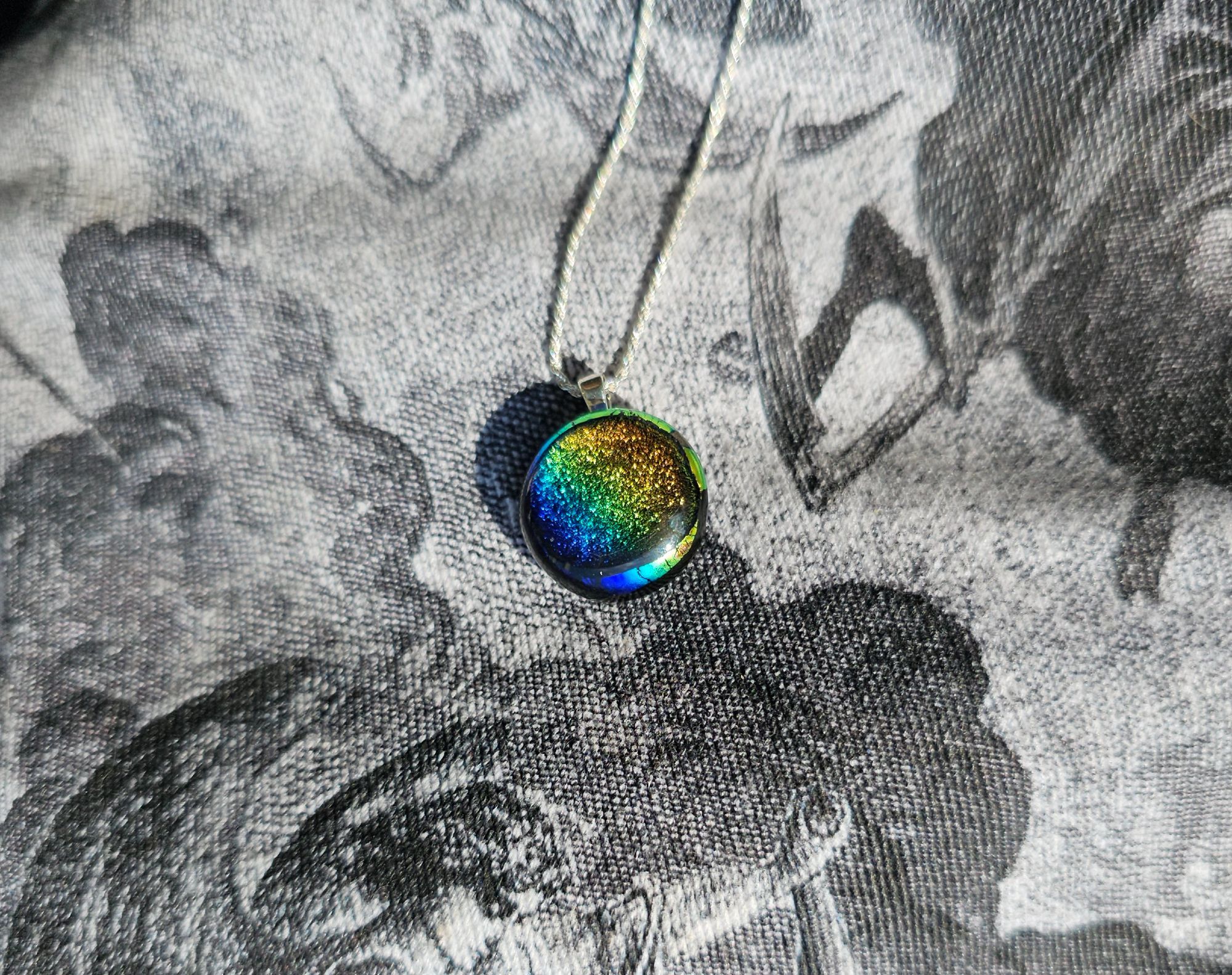 Photograph of a glass pendant on a silver chain from Nora.zone

The pendant is rainbow colored, round, about the size of a US penny, and is made of dichroic glass. The background fabric, with black and white depictions of angels