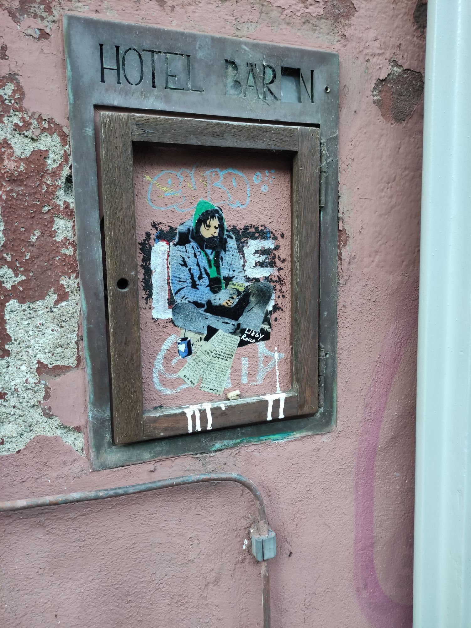 Foto von einem Graffiti in einem kleinen Schaukasten an einer Wand.
Dargestellt ist eine sitzende, scheinbar Obdachlose Person die etwas Geld sammelt. Sie trägt eine blaue Jeansjacke uns Hose, schwarze Sneaker und einen grünen Kapuzenpullover sowie einen schwarzen Schal oder Tuch. Die Kapuze ist über den Kopf gezogen um vor Kälte zu schützen. Vor der Person ist ein eingerissener Zeitungsausschnitt geklebt. Es ist eine Annonce zur Wohnungssuche.