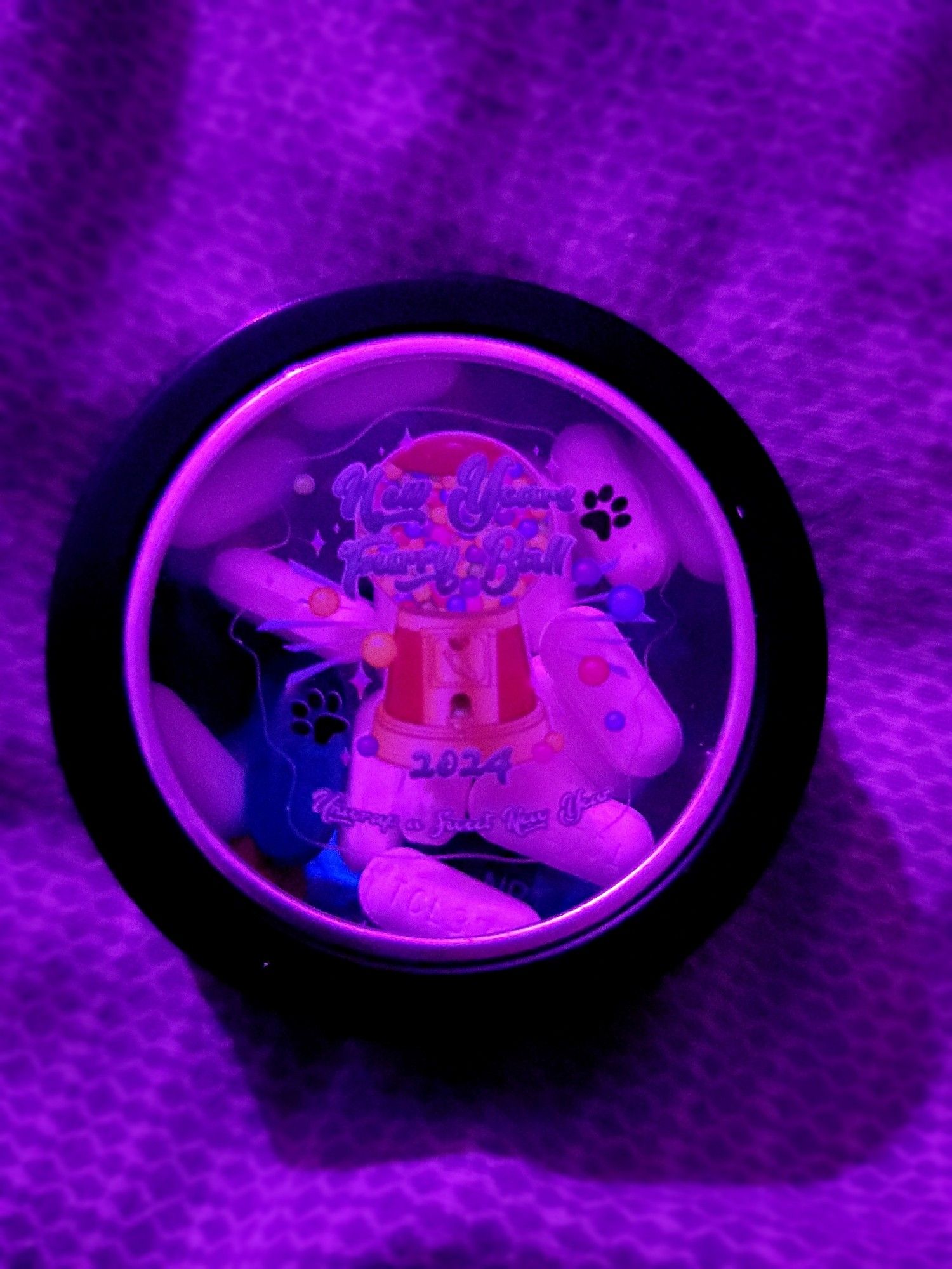 A small round tin with a clear window in the lid, through which are visible pain reliever capsules. The sticker on the window shows a gumball machine with paw prints, confetti, and the legend "New Years Furry Ball 2024 - Unwrap a Sweet New Year"