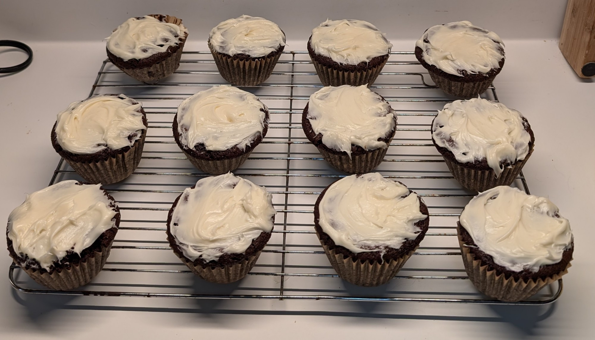 A dozen chocolate cupcakes topped with vanilla icing, cooling on top of a wire rack 