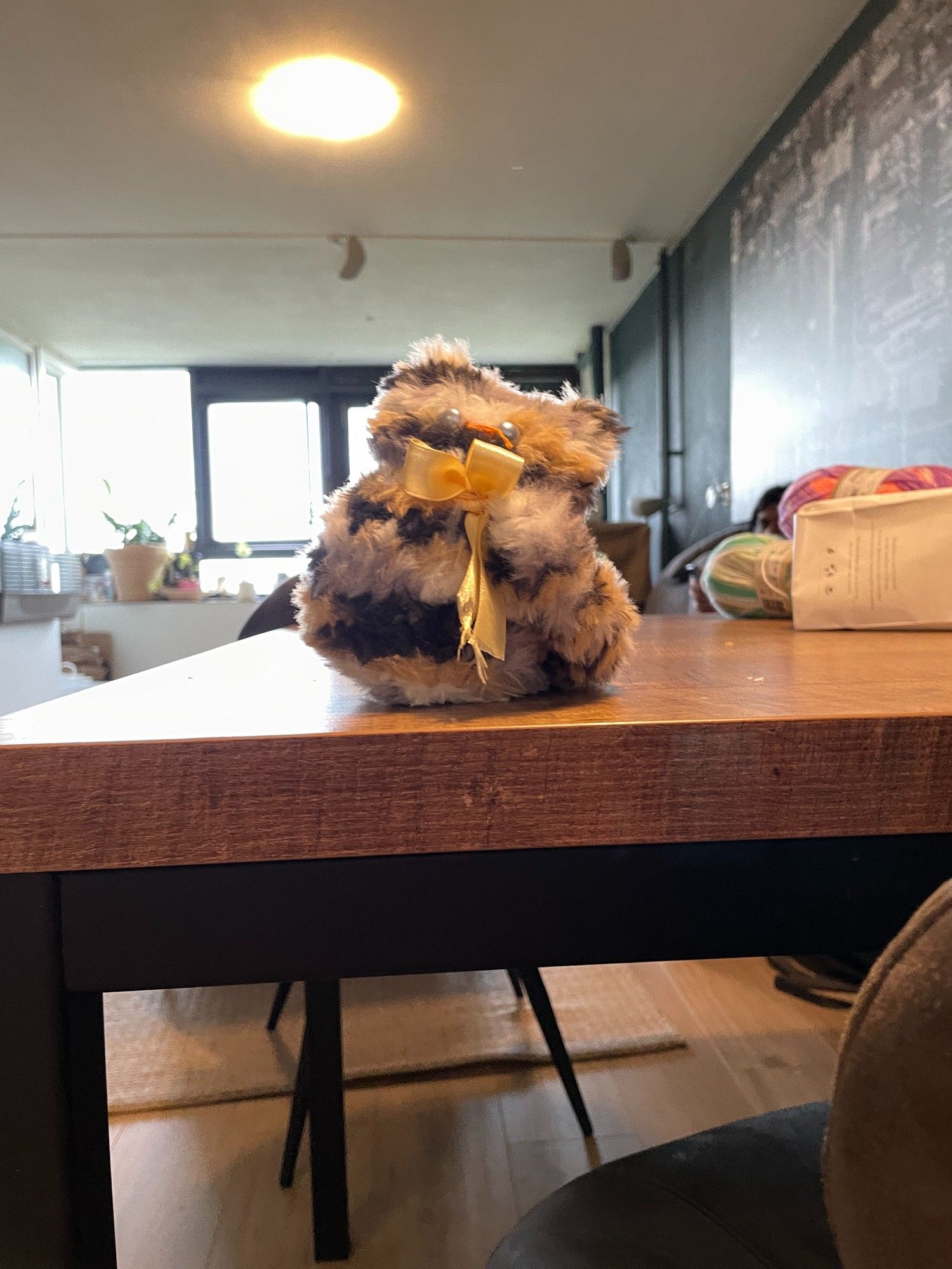 A super-chubby, fluffy cat-plushie, sitting on a wooden table. It's knitted with a yarn in varied shades of brown and white fur, giving it a textured look. Its eyes are large and round buttons with orange and black coloring, giving it a quite owlish appearance. There's a pale yellow ribbon tied into a bow around the kitty's neck. A thick, fluffy tail is curling around and up its left side. Its head is slightly tilted sidewards, so it looks kind of curious. The model is a one-piece knit and has no legs.