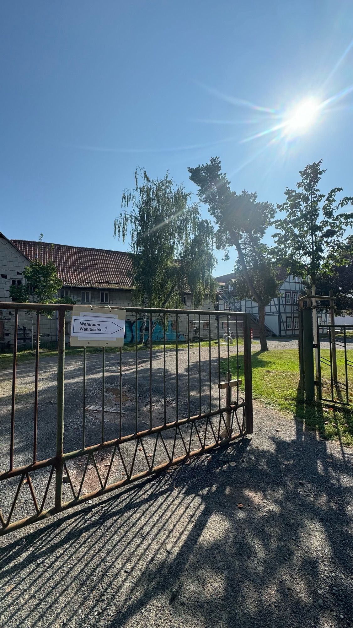 Das Bild zeigt ein offenes Tor aus Metall, durch das man auf einen Hof blicken kann. Der Hof ist von einem älteren Gebäude mit Ziegeldach und einem Fachwerkhaus umgeben. Im Hintergrund steht ein großer Baum. Im Vordergrund ist das Schild "Wahlraum Wahlbezirk 9" am Tor befestigt. Die Sonne scheint hell am klaren blauen Himmel und wirft deutliche Schatten des Tors und der Bäume auf den Boden. Es handelt sich um den Zugang zu einem Wahllokal.