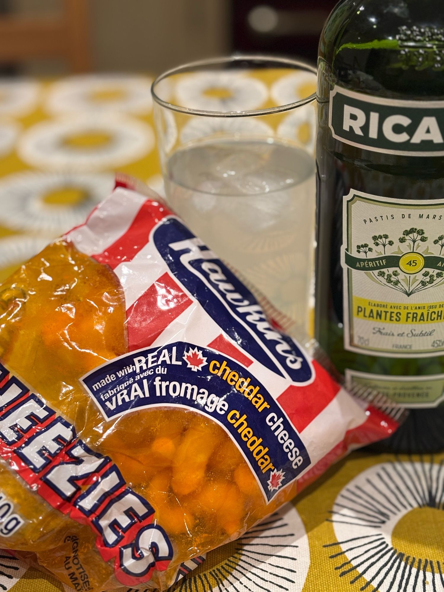 A glass of pastis behind a bag of cheezies and a bottle of Ricard