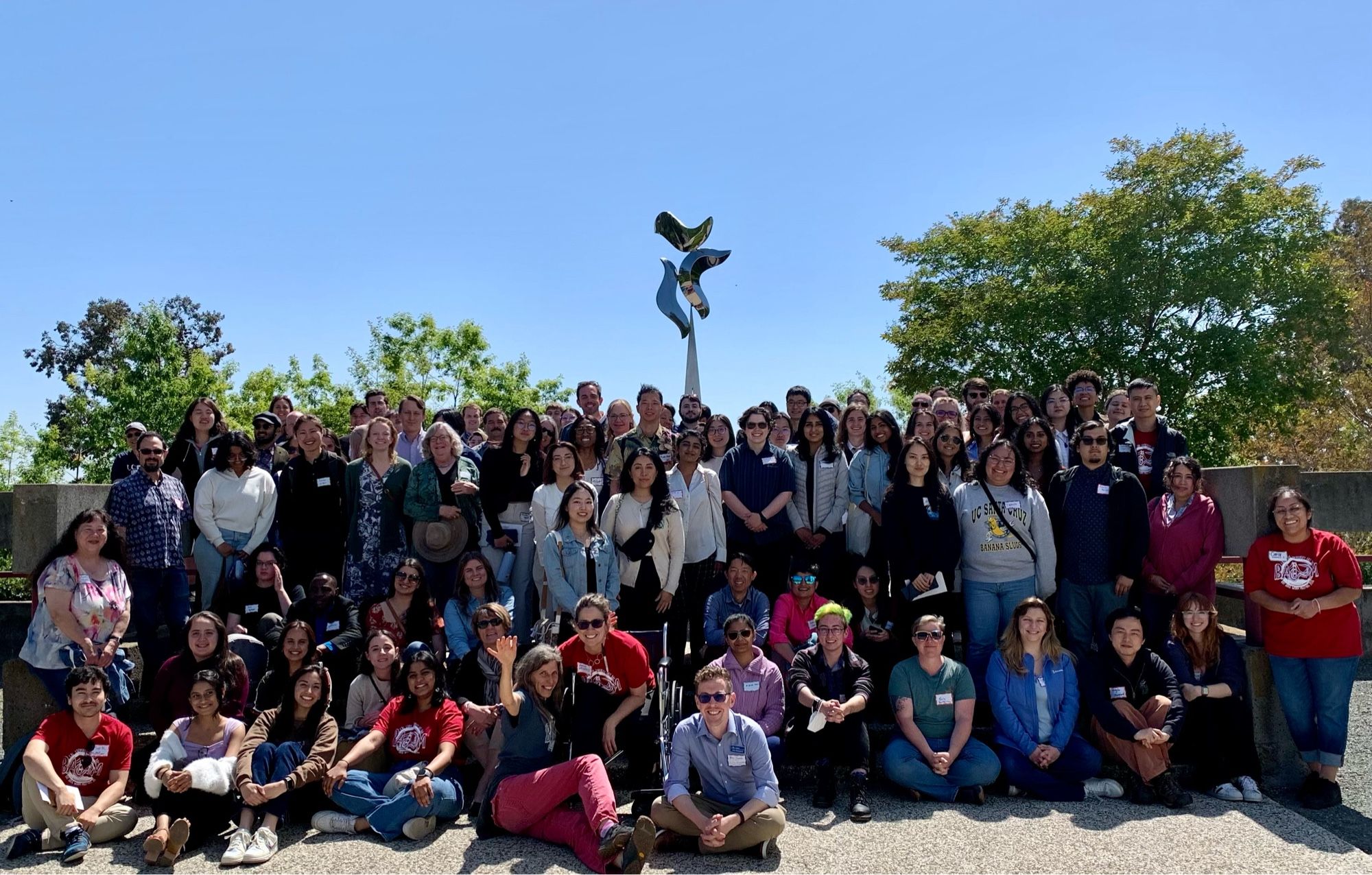 The participants of the 2024 BAWM - a group of C elegans researches seated and standing amassed under Cal State East Bay’s nictating dumpy uncs. They reach into an azure sky…Professor Maria Elena Gallegos, the organizer extrodrinare in her wheelchair sits in front.