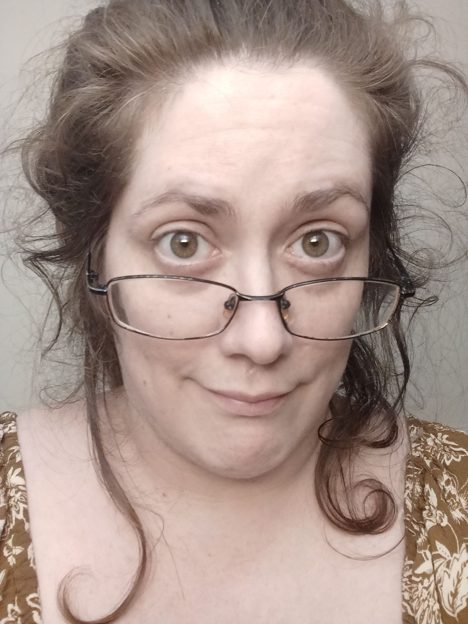 Selfie of me with my hair up very messily, with loose curls falling to my shoulders. My skin is pale, my hair and eyes brown, I've got glasses on the bridge of my nose and my dress is a light brown with a pale leafy pattern.