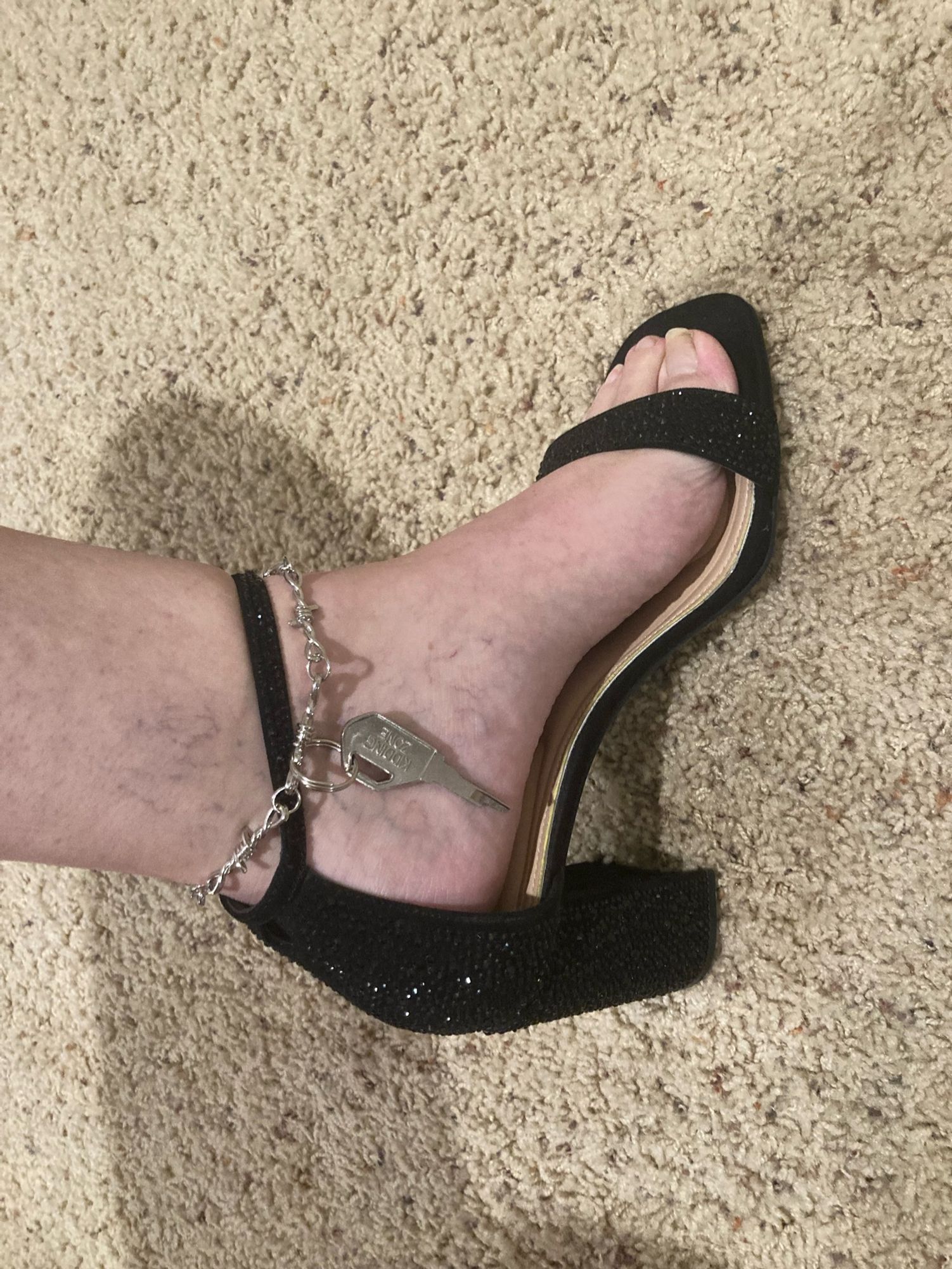 A picture of my left foot in heels, a barbed wire anklet on with a chastity key dangling from it