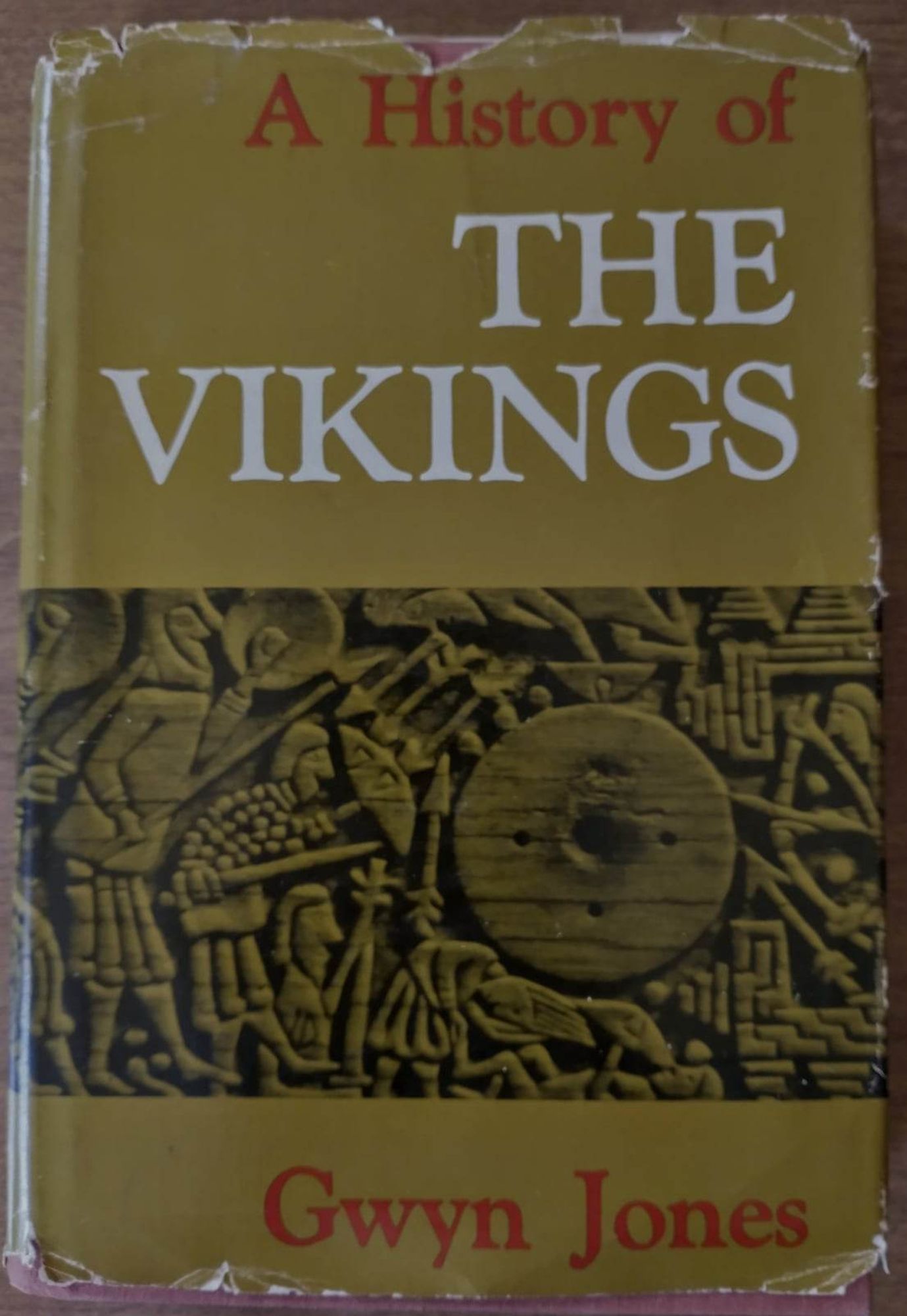 A hardcover vintage copy with dustjacket of 'A History of the Vikings' by Gwyn Jones. The cover is a plate of men with swords and other implements fashioned around a center circle. It is a woodcut. The Wheel has an arrow pointing down to it, there is one man lying prone beneath it. It's off center to the right, with several men to the left in varying height with sowrds and 2 men on the right with bow/arrow.