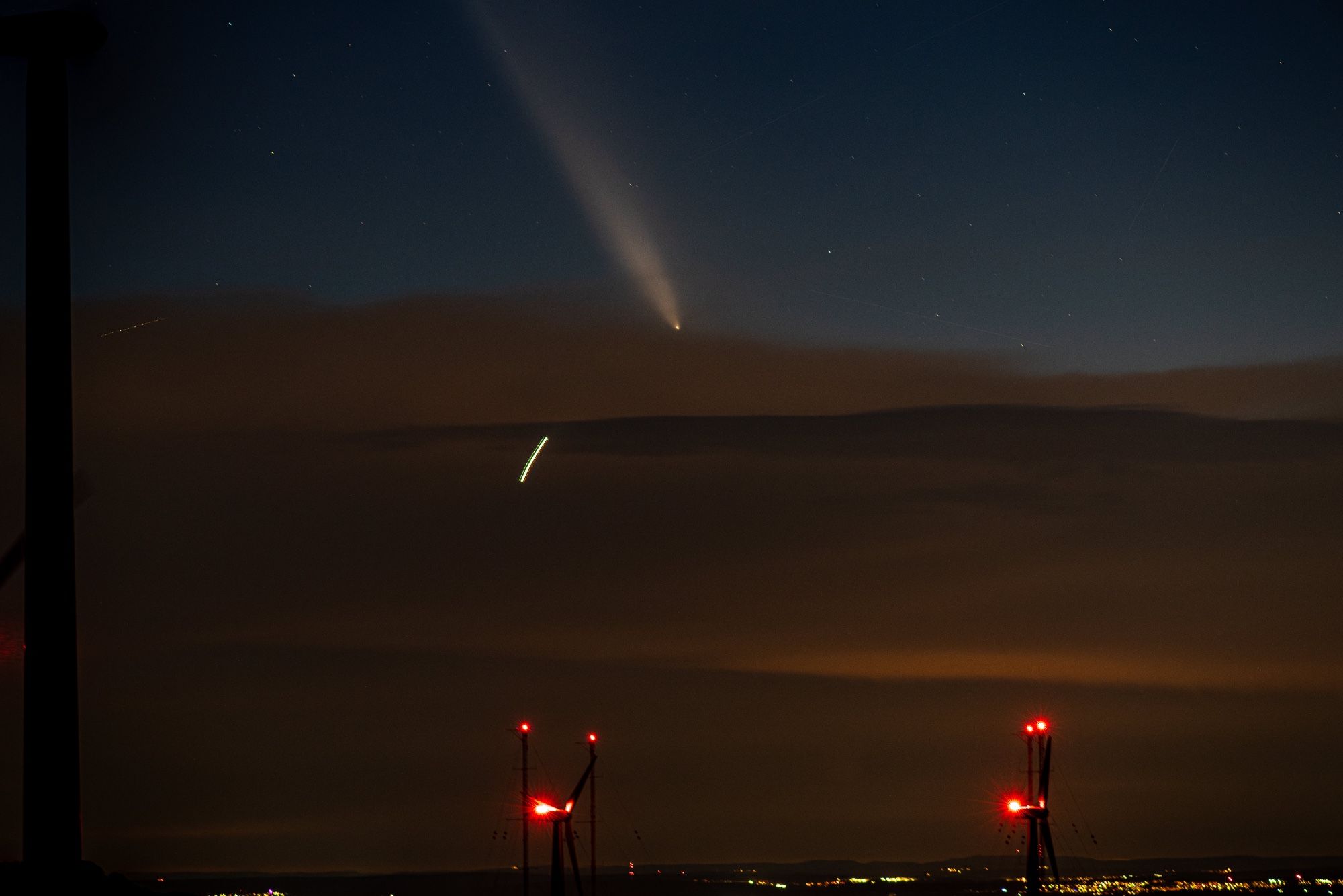 Foto nach Sonnenuntergang. Im Vordergrund Windräder und Hochantennen mit roten Signallampen, unscharf sind Lichter von Ortschaften zu erkennen. Am Horizont eine Wolkenfront, ein Drittel ist Wolken los, da ist der Komet zu sehen, neben Sternen und mehreren Satelliten Spuren. Im Bereich der Wolken die helle Spur eines Flugzeugs.