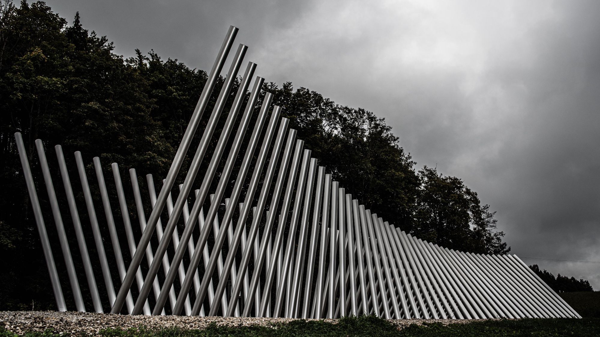 Eine Skulptur aus 2 Reihen Röhren, deren Neigung sich wellenförmig ändert, entstehen neu Muster je nach Betrachtungswinkel. Die Höhe der Röhren beträgt mehrere Meter, der Himmel ist stark bewölkt.