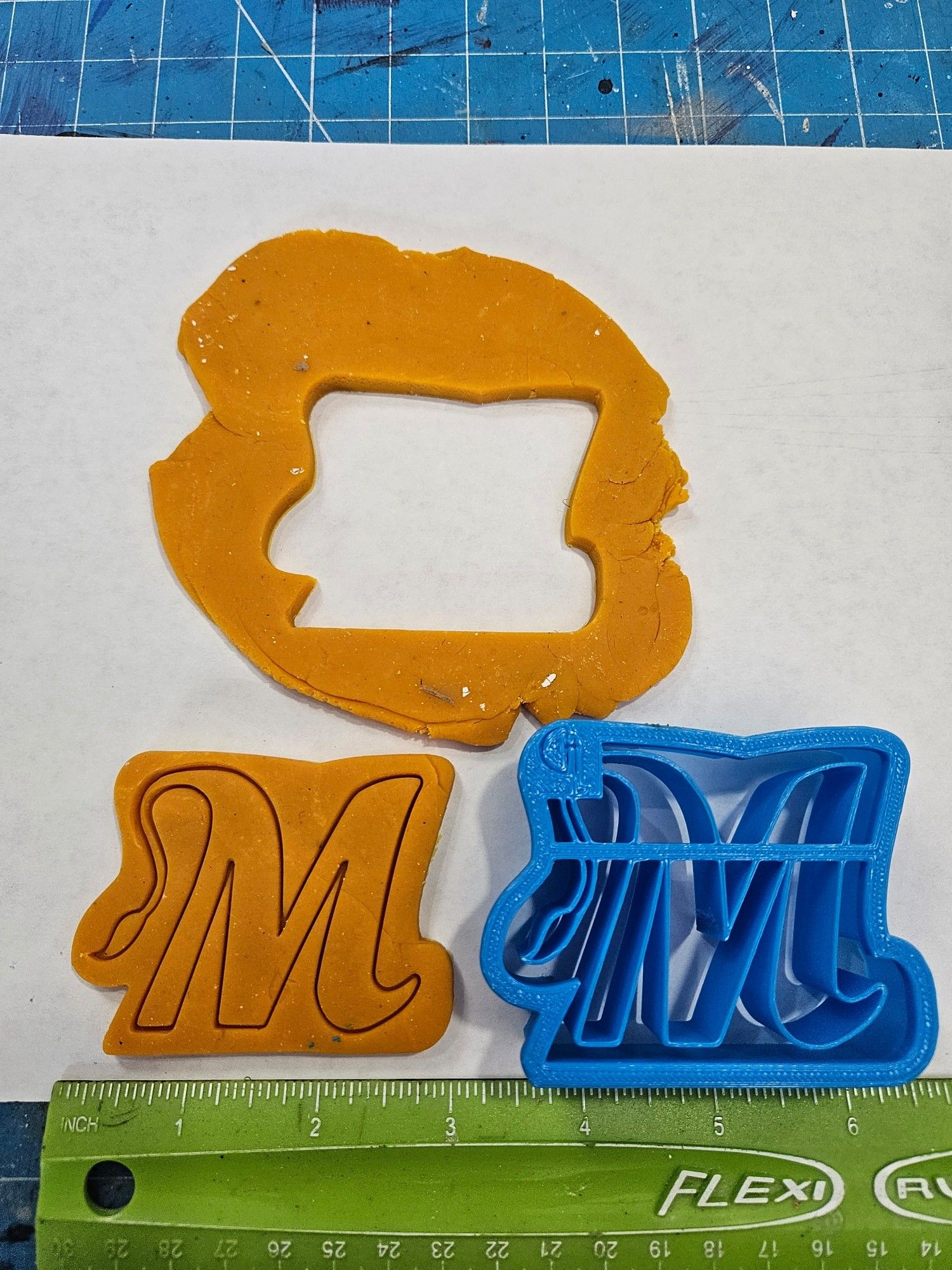 Photo of example cookie cutter that was Custom modeled and then 3D printed.   There is a rolled out patch of play doh with the cut out from the cookie, the cutter itself and the cookie cut from the play doh which shoes an imprint on its surface with a stylized letter M