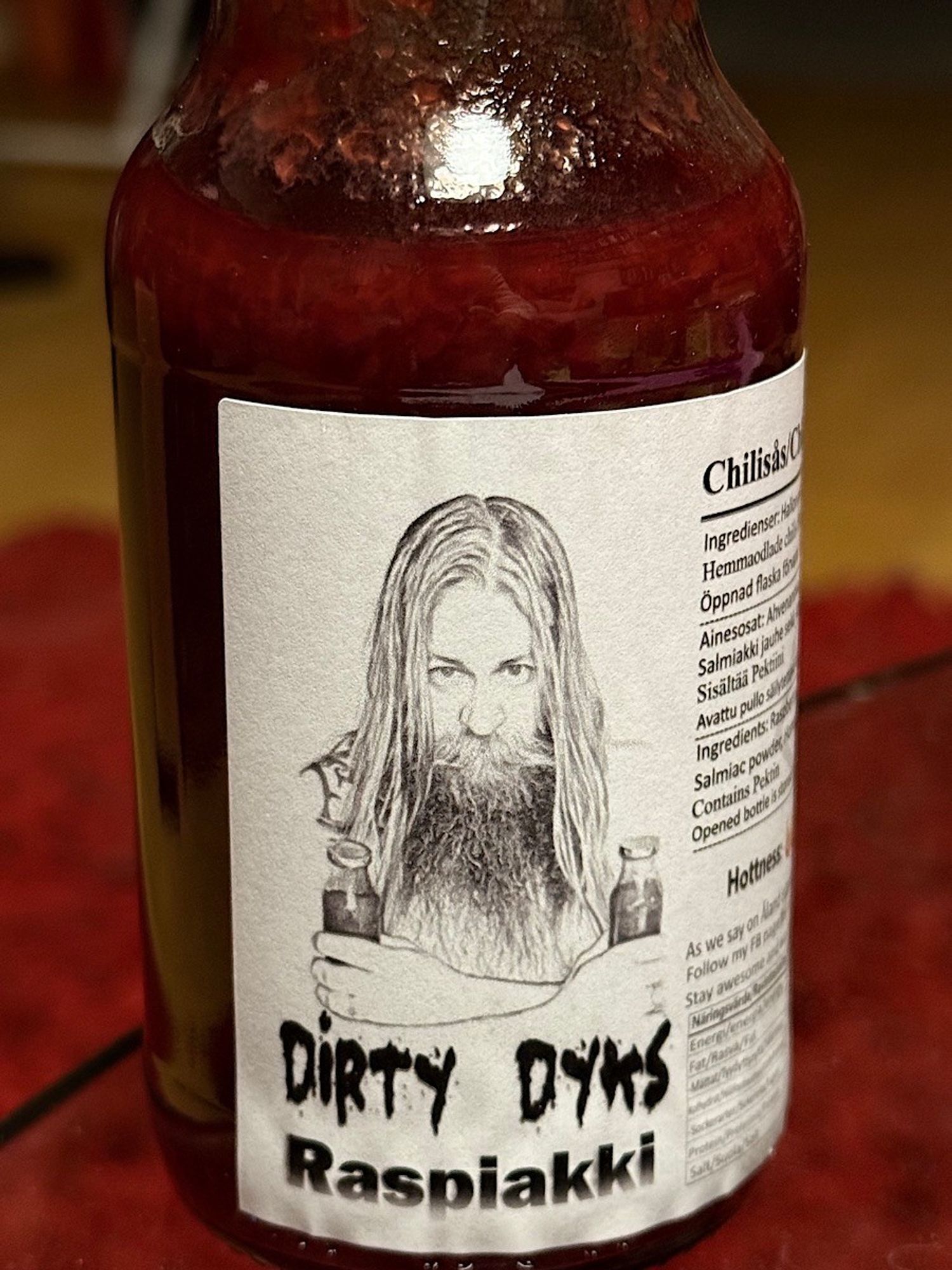 A glass bottle of chili sauce labeled "Dirty Dyks Raspiakki." The label features a black-and-white illustration of a man with long hair and a beard, holding two bottles. The design includes ingredients and product details in Swedish. The sauce has a deep red color that looks absolutely delicious.