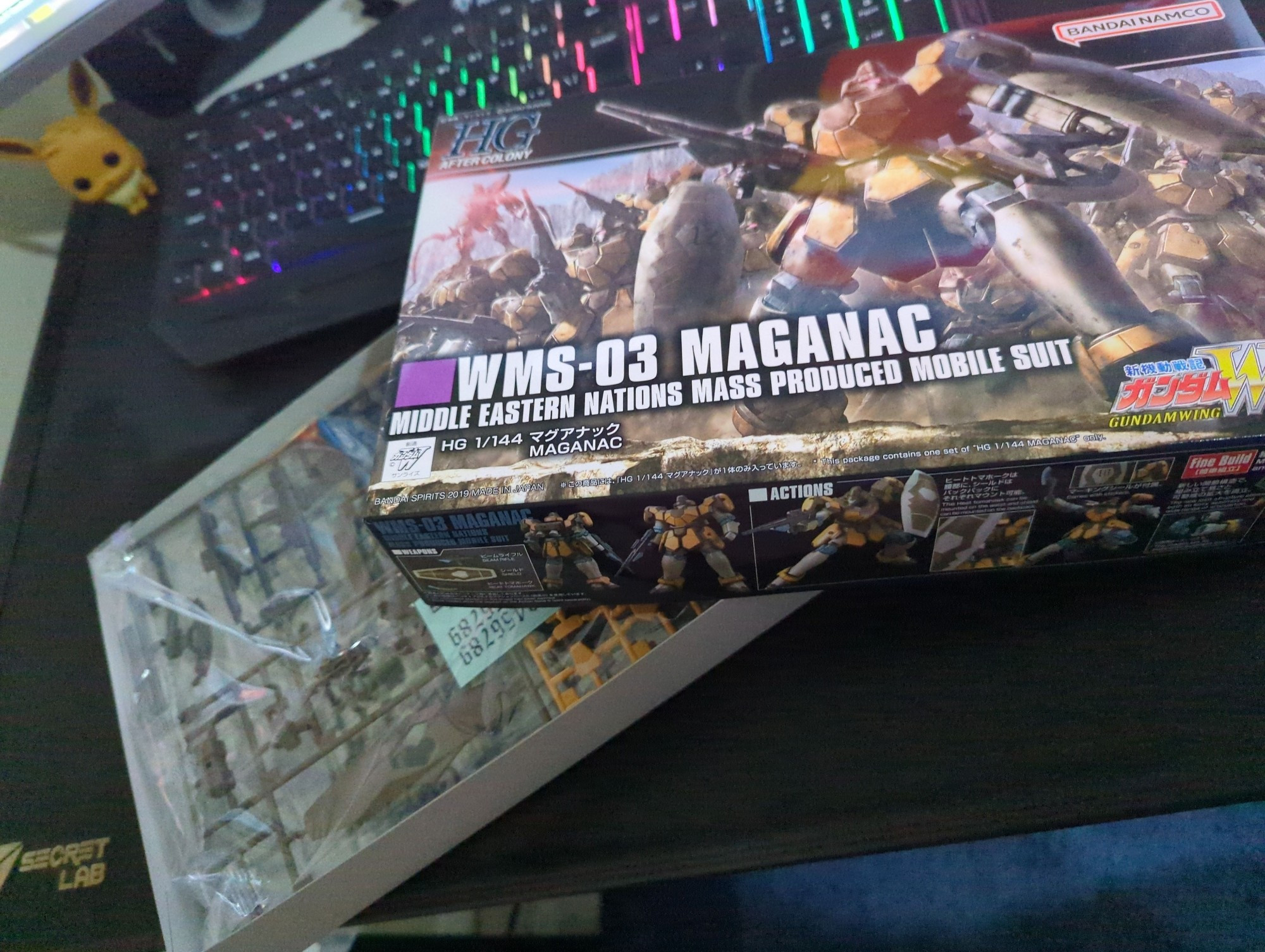 An open box of the HGAC Maganac gunpla kit showing the front of the box and the still bagged rails in front of a gamer keyboard. An Eevee Funko pop can be seen on the background