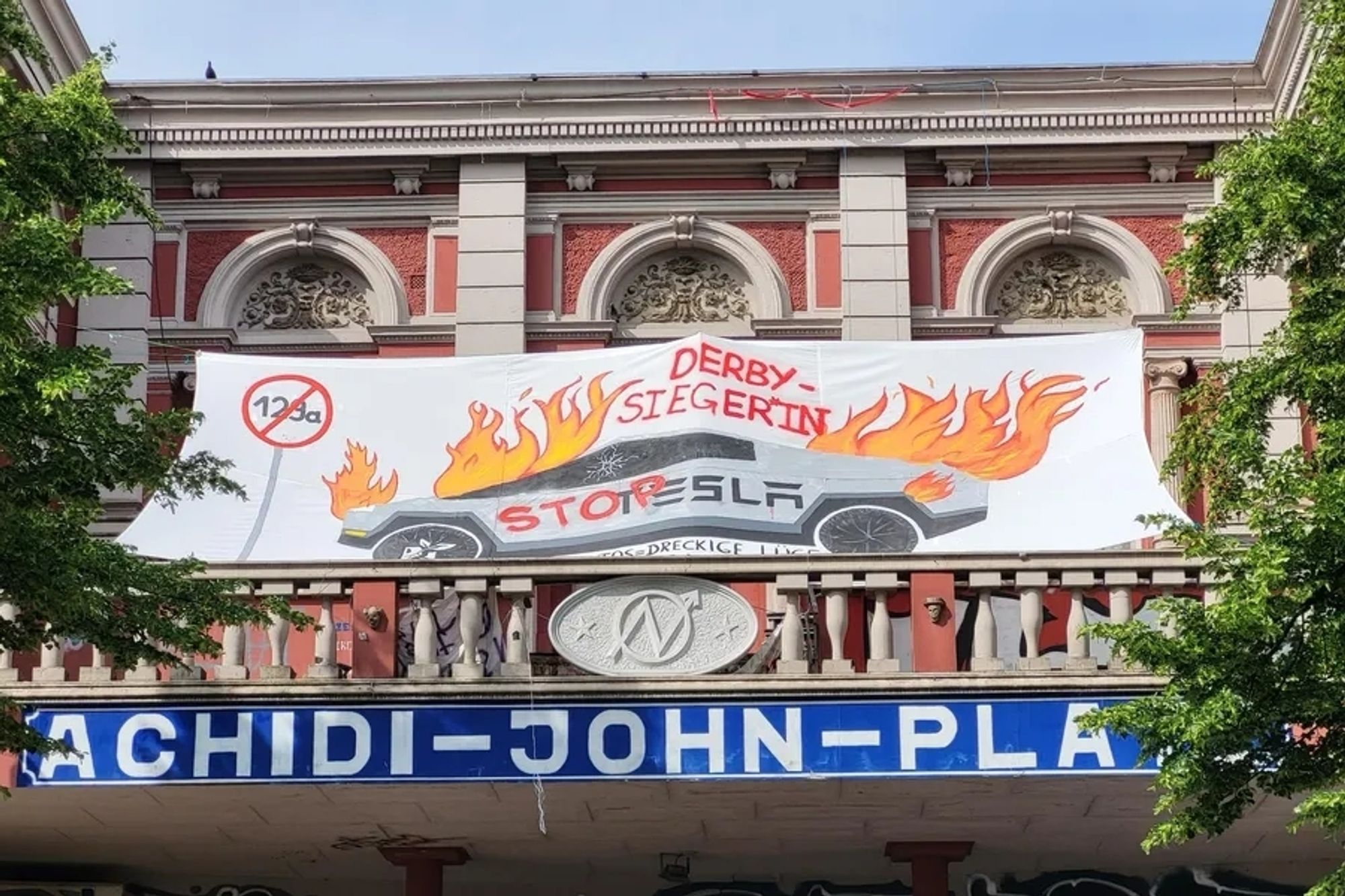 Balkon der Roten Flora in Hamburg. Auf diesem ein Plakat mit einem brennenden Tesla Cybertruck. Auf dem in Rot "Stop" und in grau "Tesla" steht. Darüber "Derbysieger*in" und links davon ein gemaltes Straßenschild mit einer durchgestrichenen "129a"