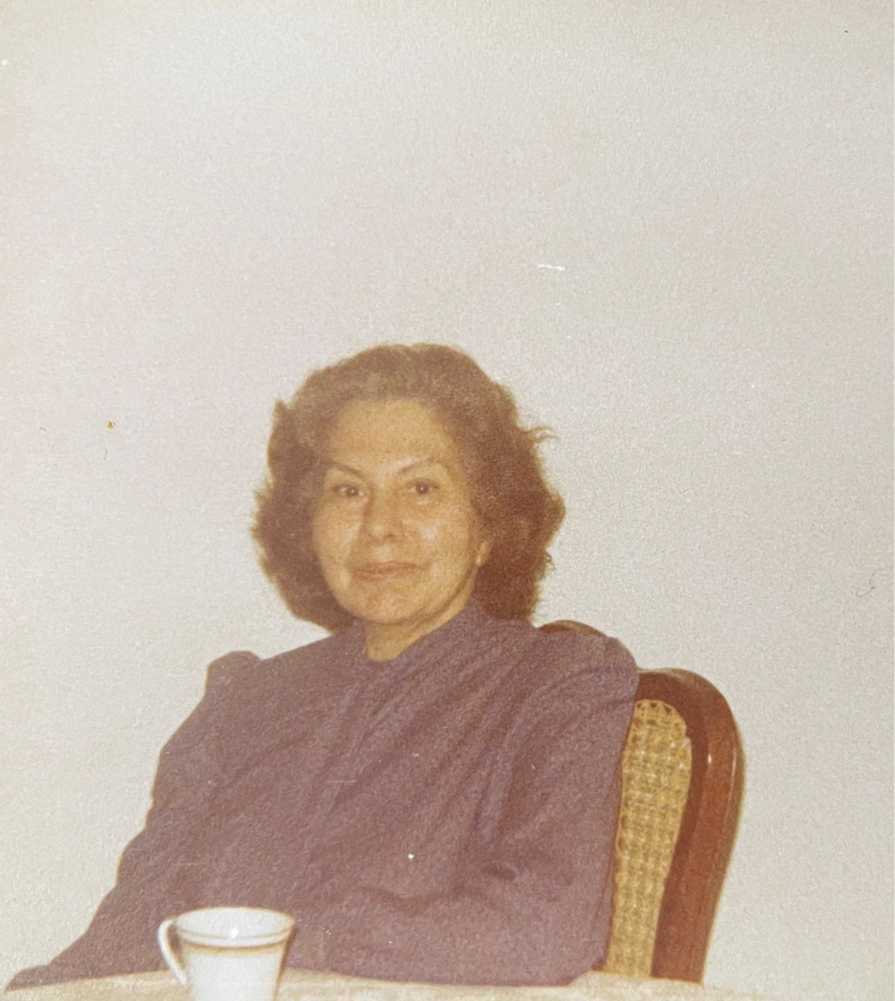 A grainy photo of an elderly woman sitting at a table. She has curly hair and gorgeous arched eyebrows.