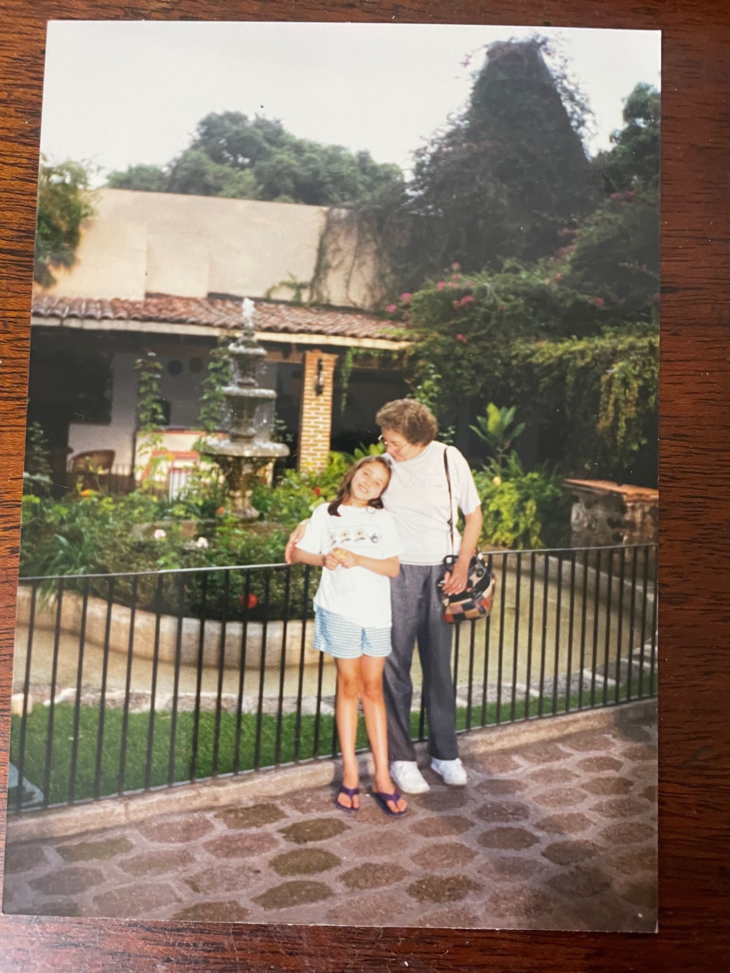 Color photo from the mid-90s. The curly-haired woman, now grey-haired, stands in the verdant courtyard of a colonial-style building with a little girl. The girl looks at the camera; the woman puts her arm around the girl and gazes down at her.