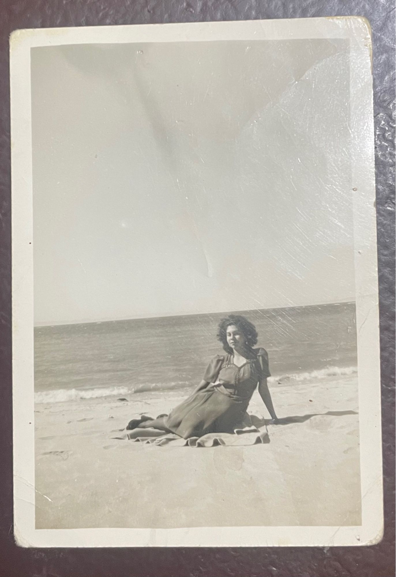 The curly-haired girl, aged 19, 1945. She sits on a blanket at the beach, leans back on one arm, and smiles mysteriously.