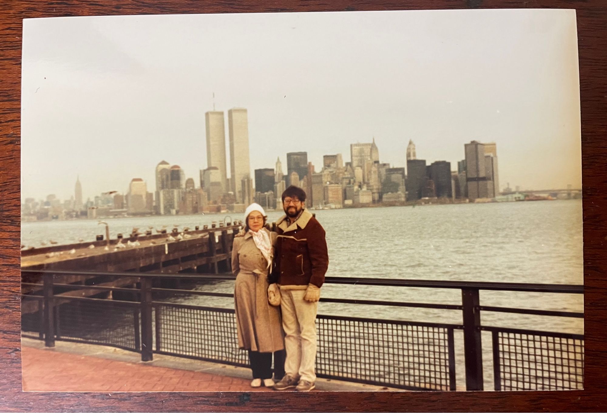 Grainy color photo: the curly haired woman poses with her oldest son on the waterfront opposite lower Manhattan. The Twin Towers loom in the background. Likely mid-1980s.