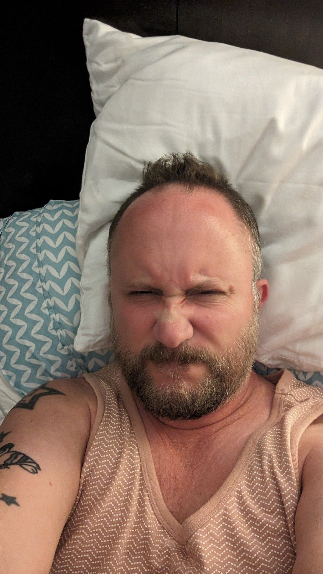 Angry gay man, lying in bed, fully clothed, narry a spare dick or hole anywhere. His ass, though not pictured, is irritated from shitting so many times in the last eight hours. He is making a face that reflects these facts.