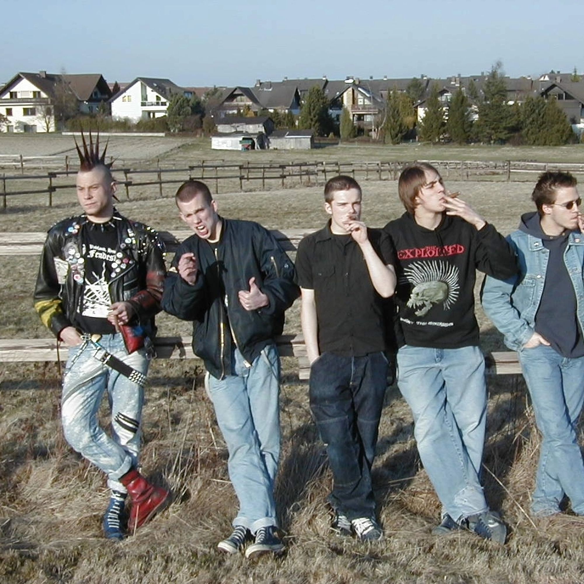 Bandfoto. Fünf junge Männer stehen an einen Weidezaun gelehnt auf einem Feld und rauchen Zigarren.