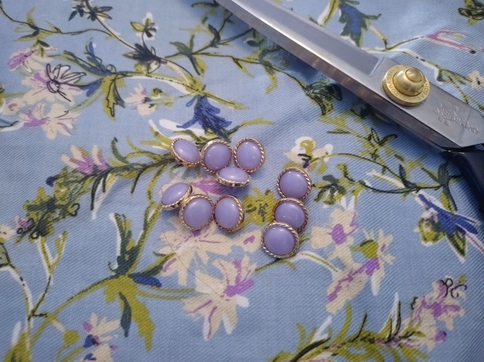 A pair of tailoring scissors, 10 lilac-coloured buttons with gold rim on a light blue fabric with white and purple flowers