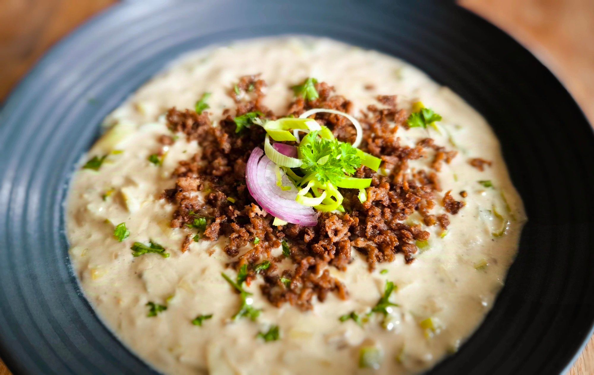 Auf einem Holztisch steht ein mattschwarzer Teller mit einer hellen, cremigen Suppe mit Lauchstûckchen. Darauf ist gebratener Krümel-Hack und Deko aus Lauch, roter Zwiebel und Petersilie.