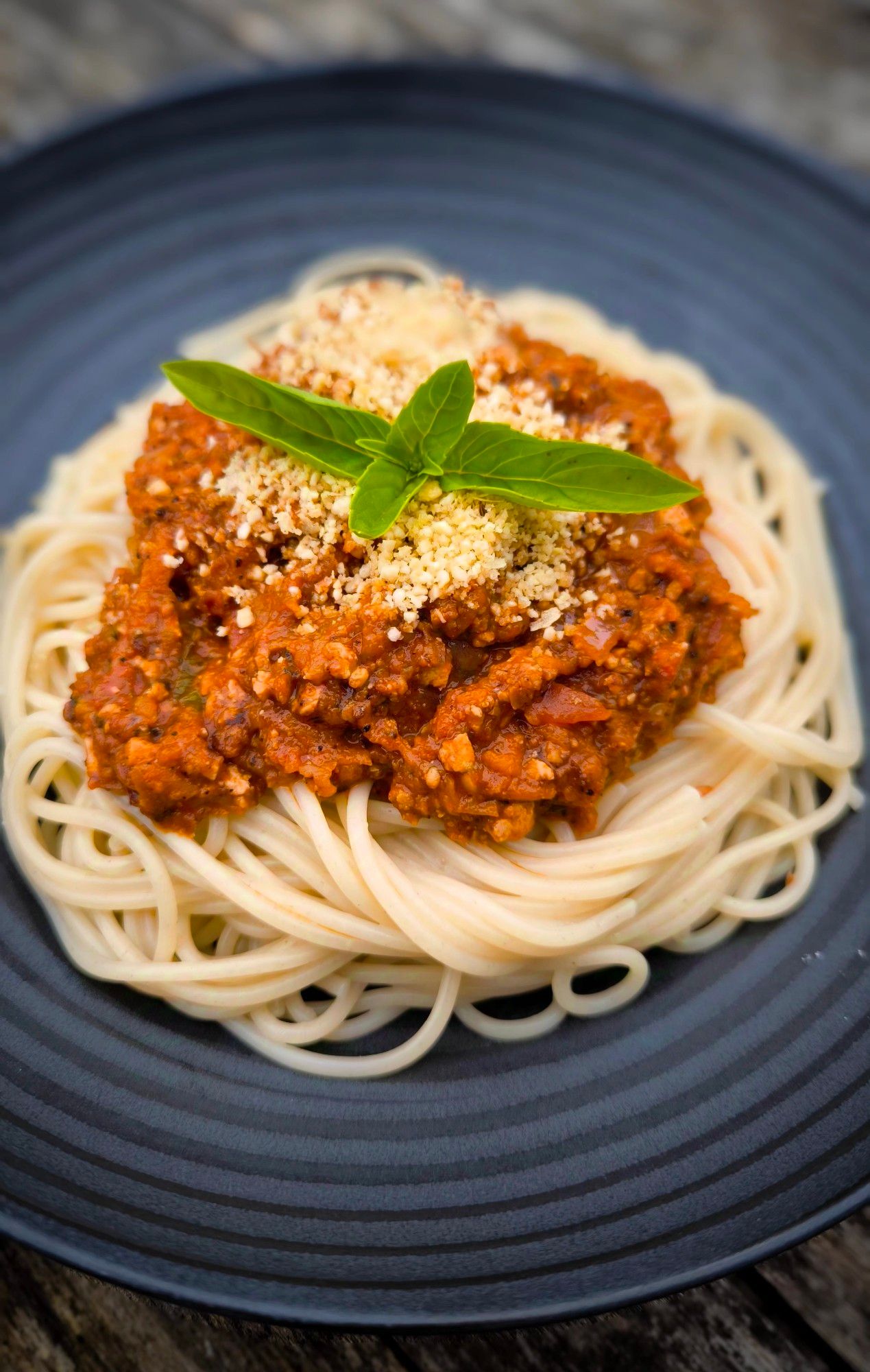 Ein mattschwarzer Teller mit einer Portion Spaghetti mit veganer Bolognese darauf. 
Dekoriert mit etwas Cashew-Parmesan und Basilikum-Blättelchen.