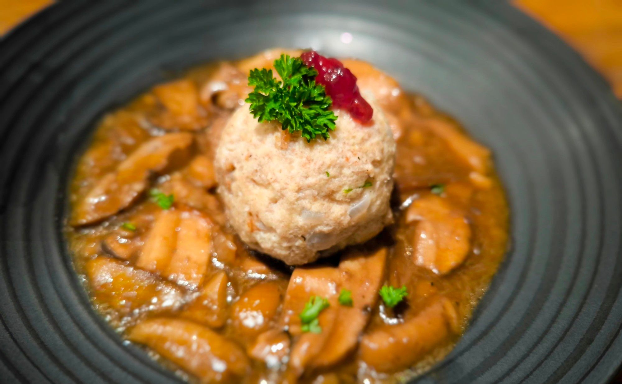 Auf einem schwarzen Teller ist angerichtet:
Braune Sauce mit Maronenröhrlingen und Steinpilzen. In der Mitte ein Semmelknödel, darauf etwas Petersilie und Preiselbeeren.