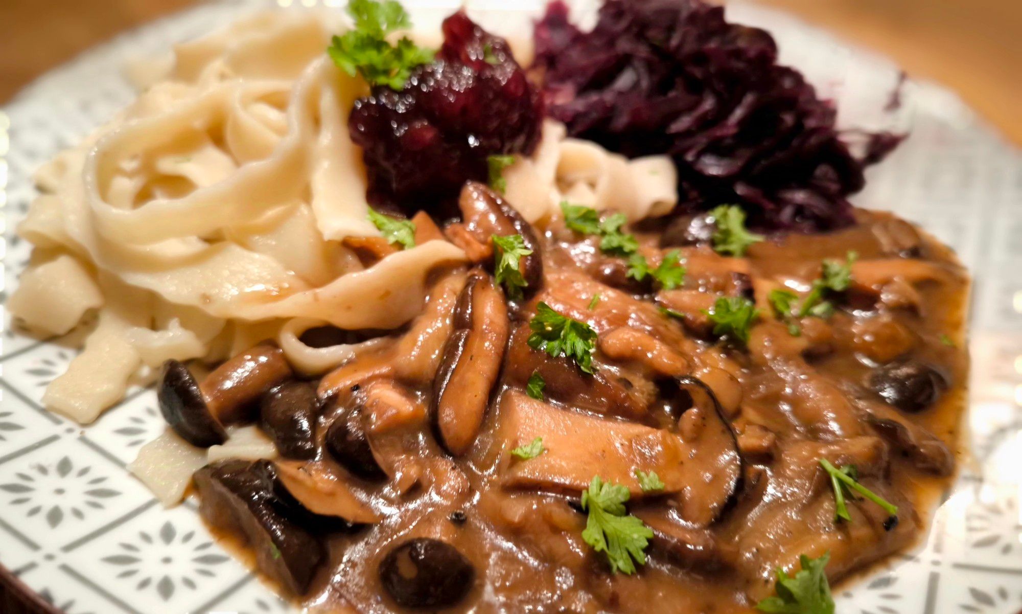 Auf einem beige gemusterten Teller ist angerichtet:
Links Bandnudeln mit gewelltem Rand, rechts hinten Rotkohl, vorne braune Waldpilzsauce mit Petersilie. In der Mitte ein Klecks Preiselbeeren.