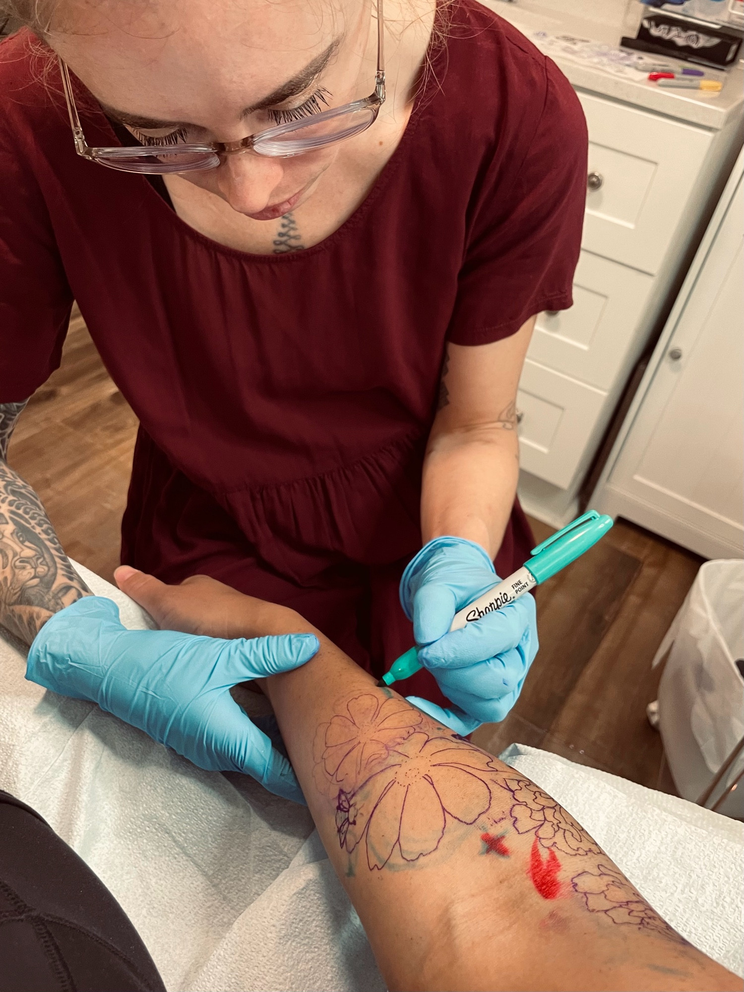 Tattoo artist Marie Reverie using a teal sharpie to draw part of a tattoo stencil on a forearm 