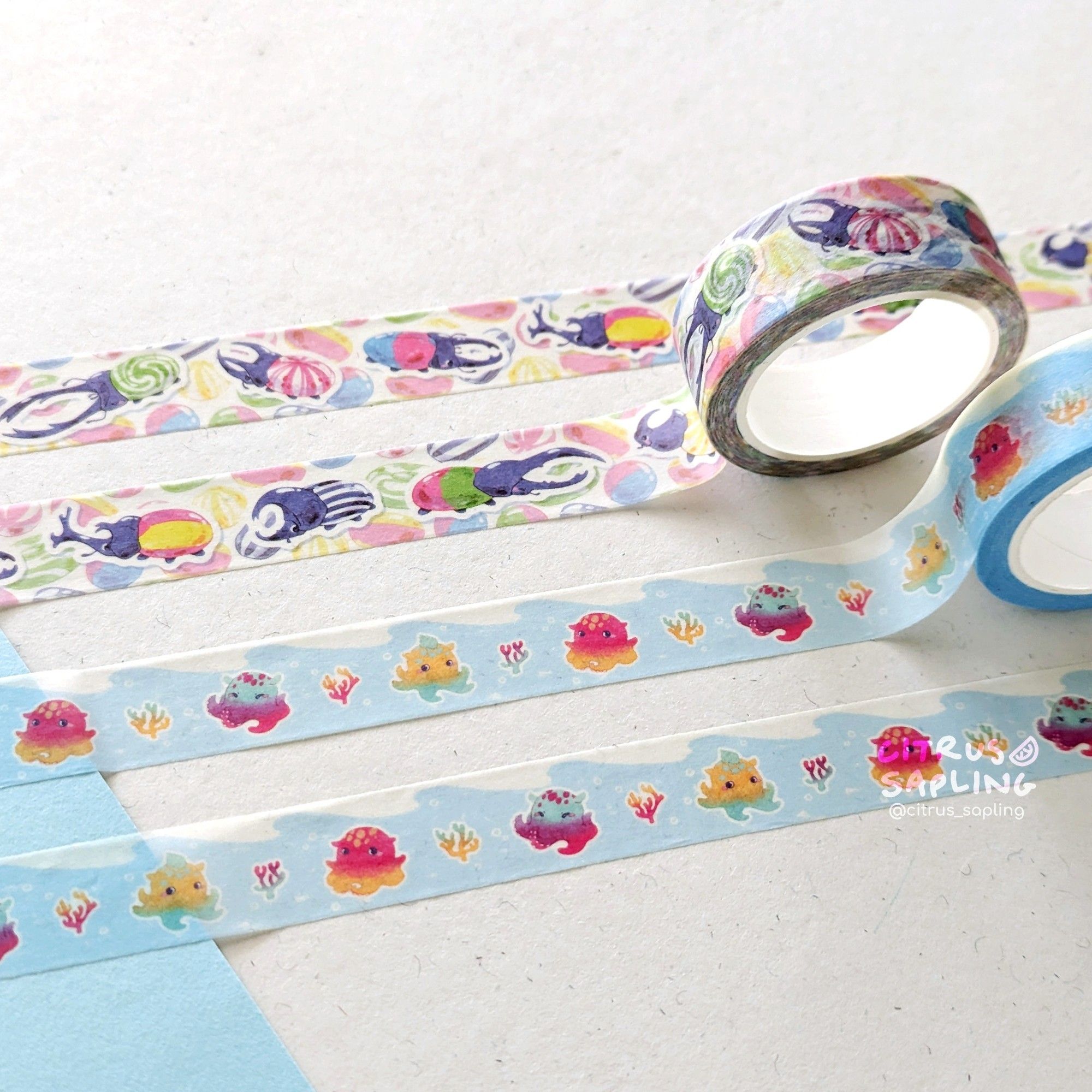 A photograph of two washi tape designs. The first one has a variety of beetles with hard sweet patterns, and more sweets in the background. The second has an alternating pattern of Dumbo octopus and coral, with an ocean wave above them.