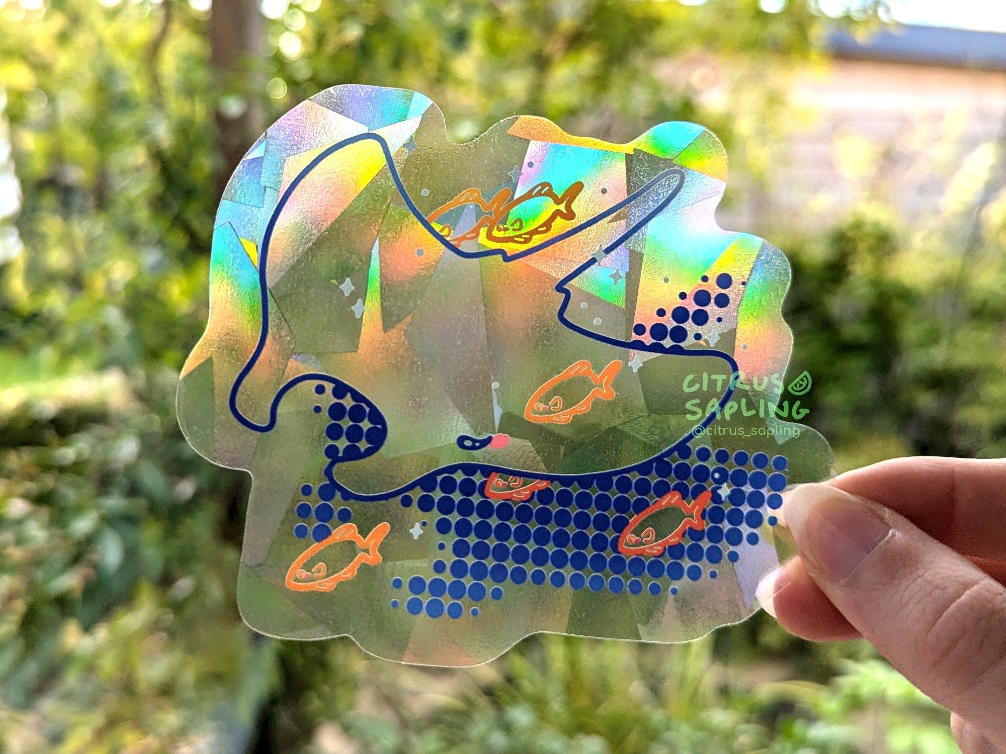 Photograph of a clear holographic suncatcher sticker against a leafy green background. The sticker features a manta ray with orange fish around it, and a halftone shadow below.