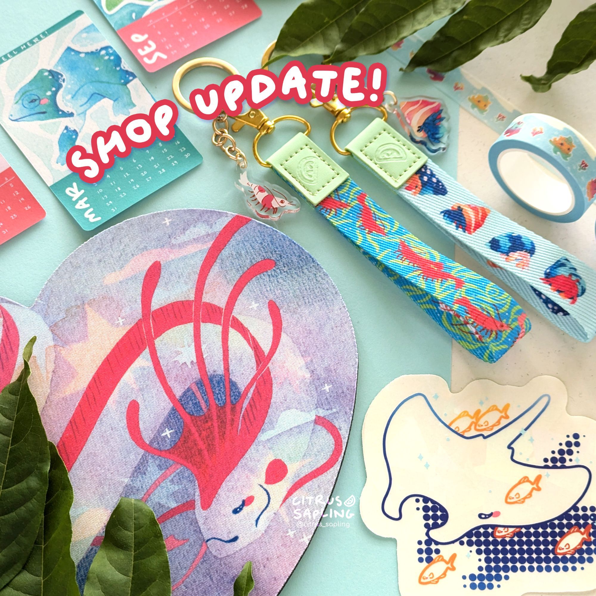 Flat lay photo of products including an oarfish mouse mat, shrimp and hermit crab lanyards, and manta ray suncatcher, framed by leaves. Text reads "shop update!".