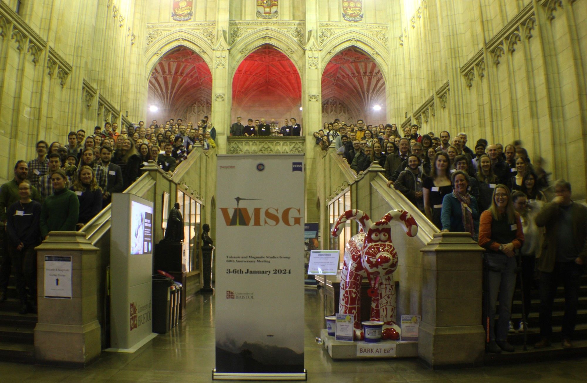 Photo of attendees at VMSG 2024. Everyone is stood up two sets of stairs which flank the entrance hall of a grand gothic building.