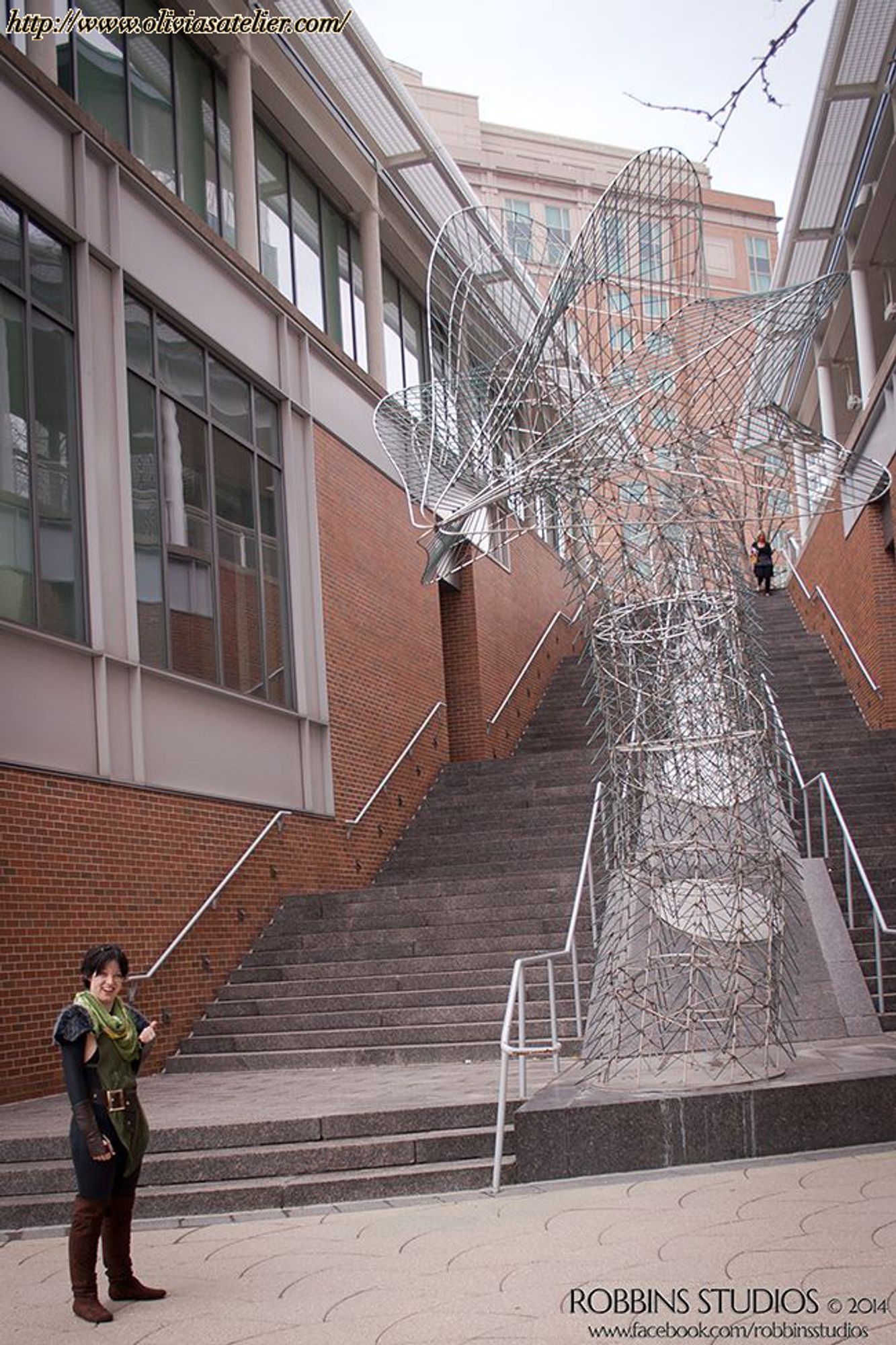 A white, non-binary person dressed as Merrill from Dragon Age 2. They are standing at the bottom of a flight of stairs, with a large metal sculpture like a dragon or serpent next to it. They are gesturing to the sculpture with their thumb.