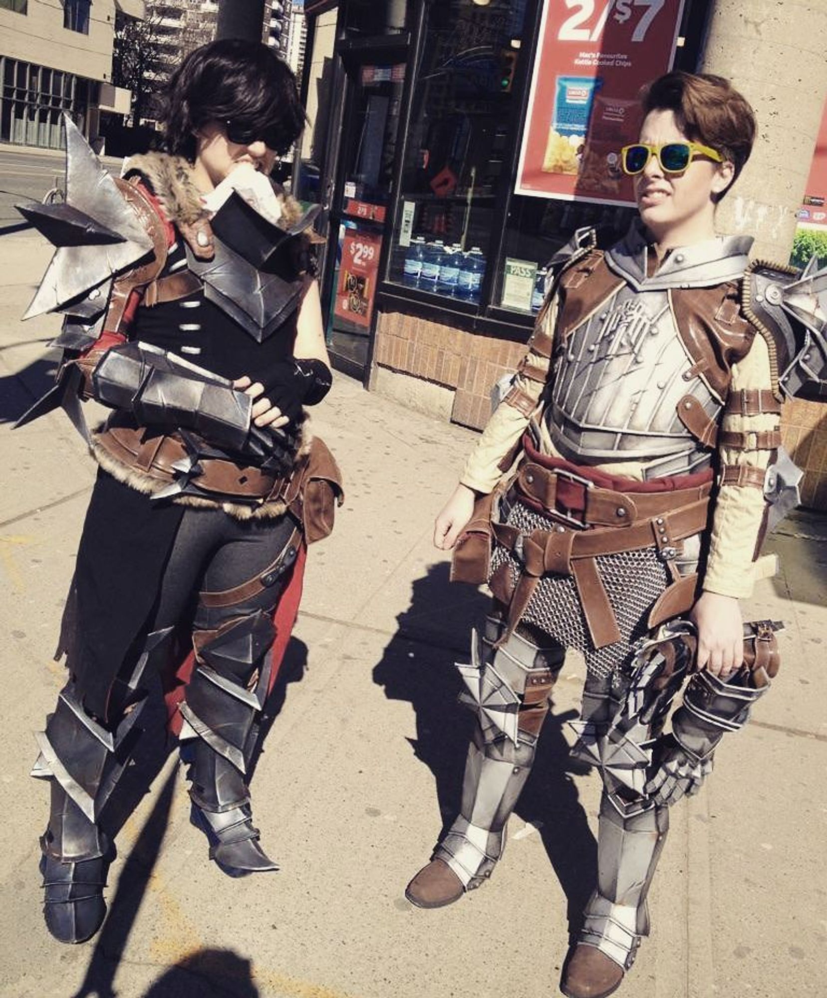 Two people dressed as Hawke from Dragon Age 2 and Krem from Dragon Age: Inquisition. They are standing on a sidewalk and wearing sunglasses. They both are making slightly uncomfortable faces.