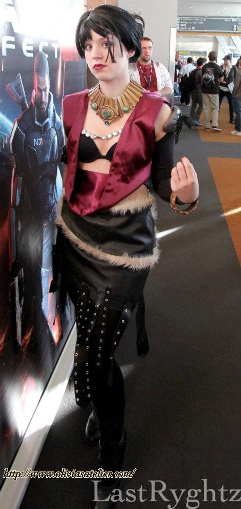 A white non-binary person dressed as Morrigan from Dragon Age: Origins. They are standing a little awkwardly, holding one hand up. There is text on the photo linking to oliviasatelier.com and the photographer's name: LastRyghtz