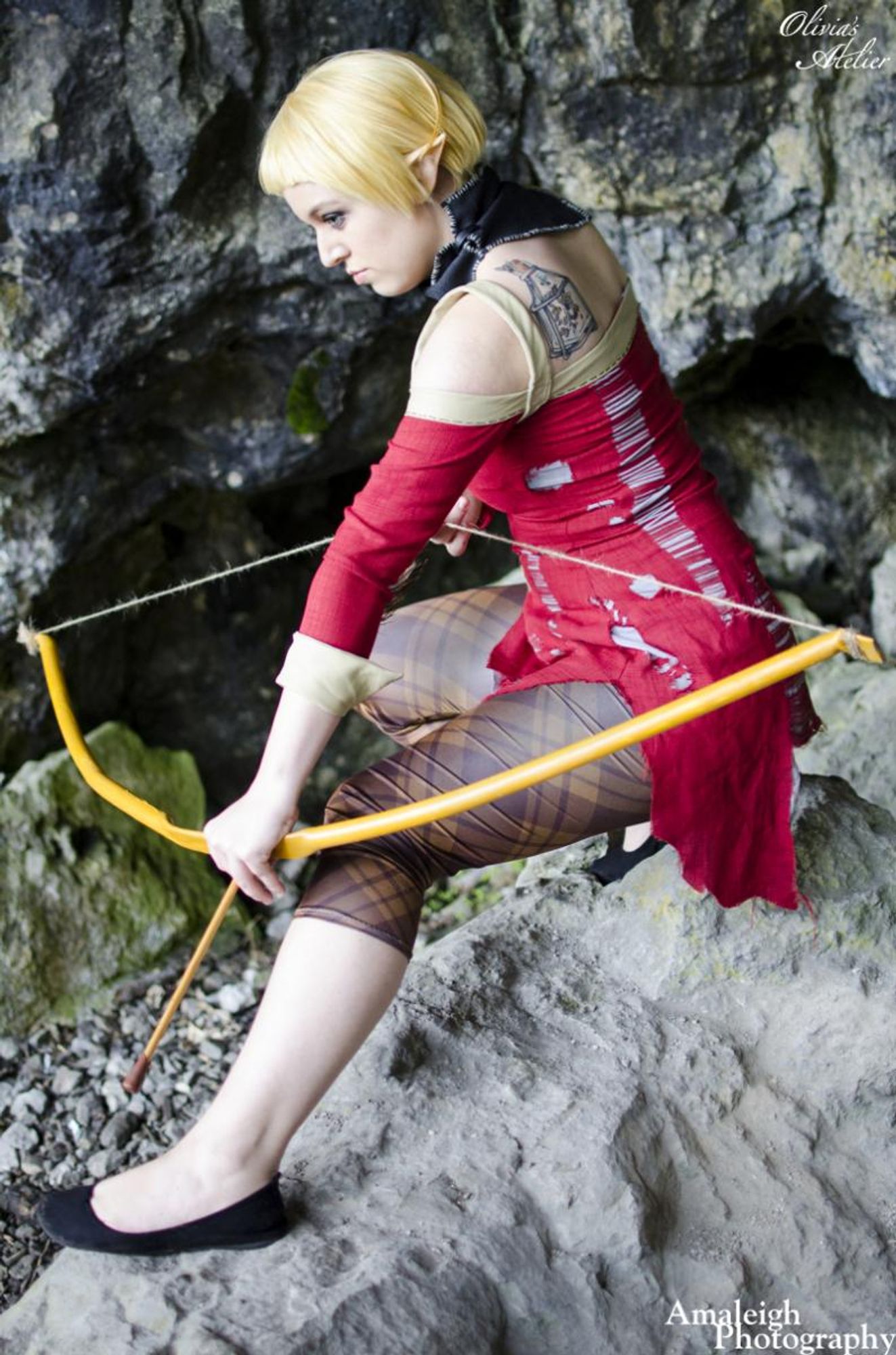 A white non-binary person dressed as Sera from Dragon Age: Inquisition. They are posed on a rock holding a bow with an arrow nocked.