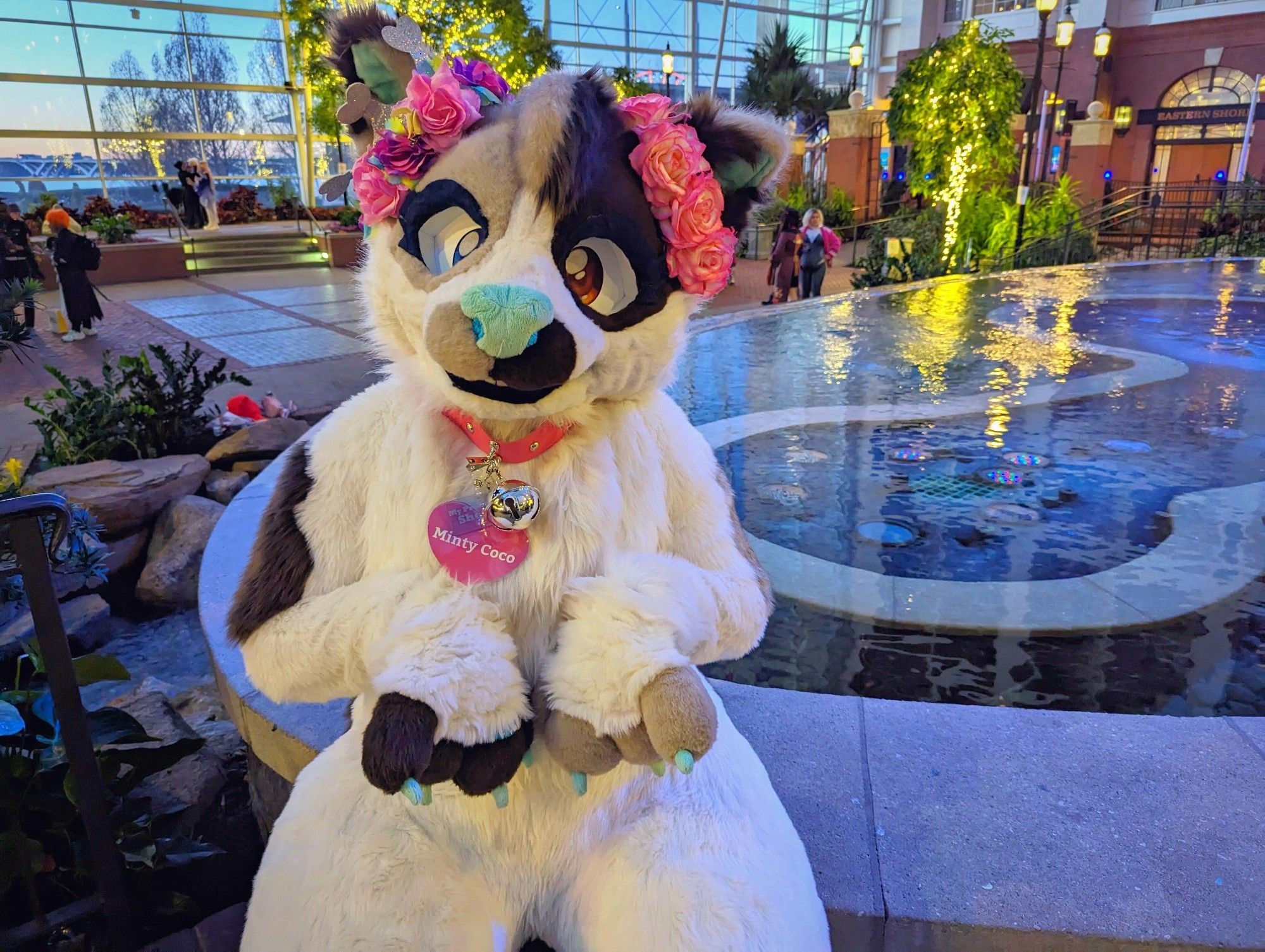 Picture of a happy cat fursuiter sitting in front of a large water fountain inside an indoor garden hotel building