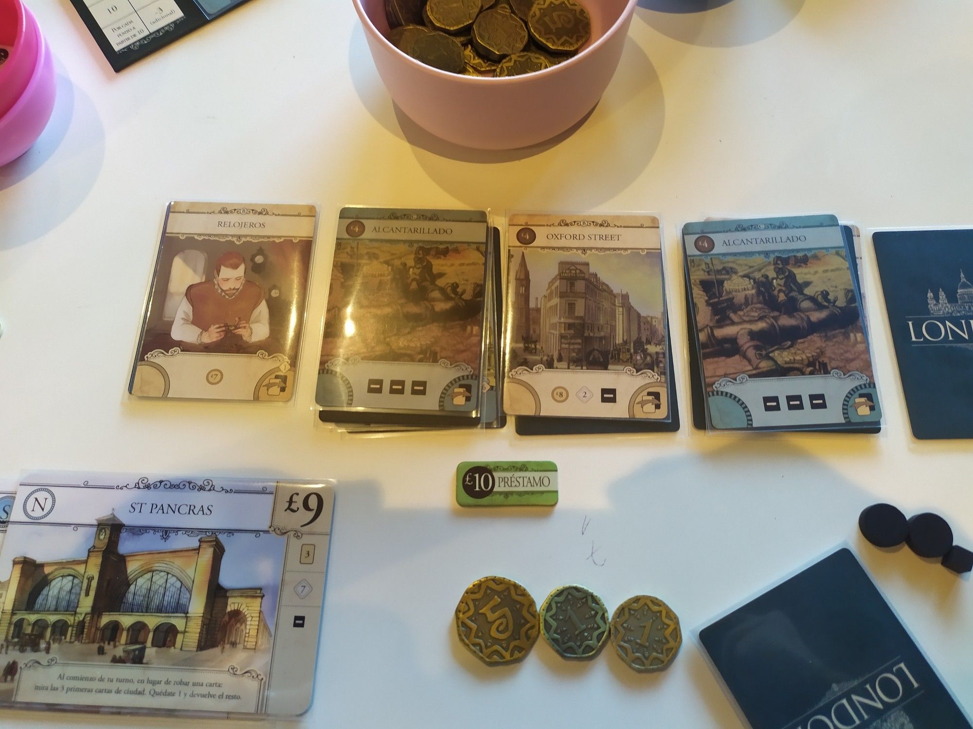 Pilas de cartas jugadas en la zona de un jugador en London, disponibles para ser ejecutadas y cobrar sus beneficios, monedas, tarjetas de distrito, un marcador de préstamo adquirido y cubitos y discos de recuento de pobreza generada por el jugador.