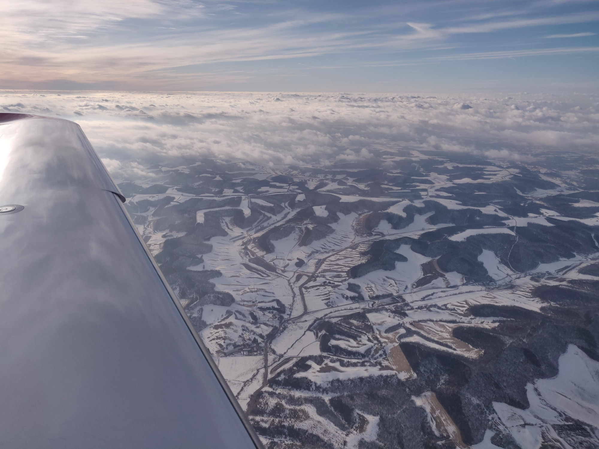 a photo taken across the wing of a small airplane looking at a snowy landscape with rolling hills and a low layer of clouds