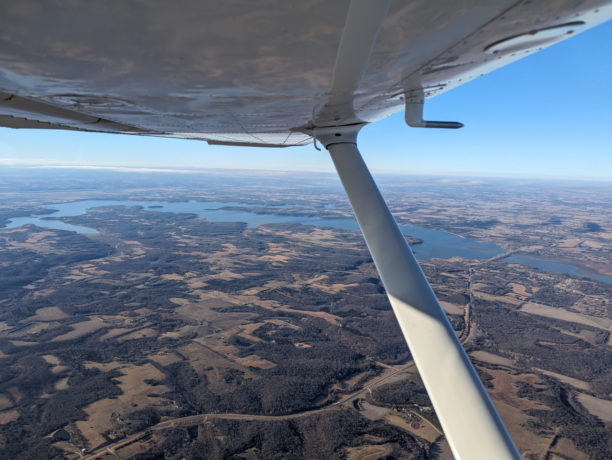 photo of taken under the wing of a small plane of a landscape with trees and farmland near a lake