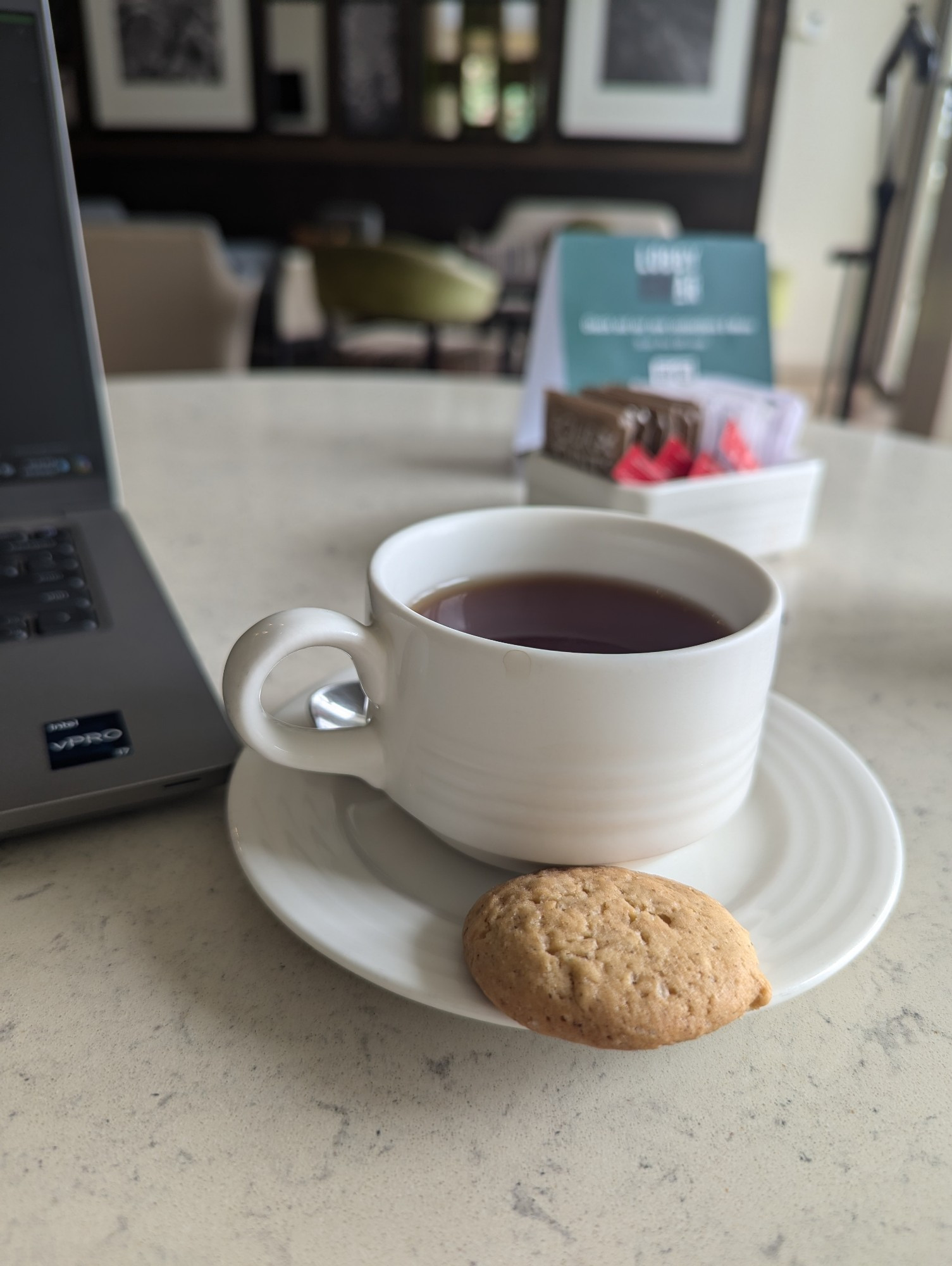 A cup of Kenyan Earl Grey tea on a saucer with a small cookie. In the corner is a NIPR computer, stuck waiting for updates. 