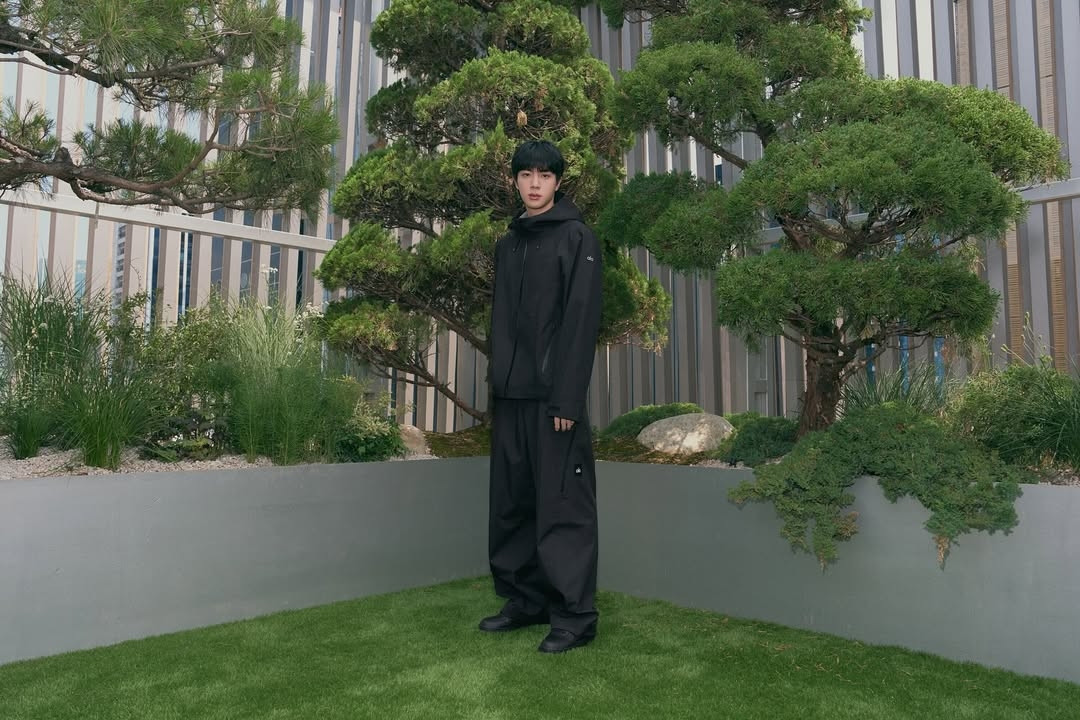 Photo of Jin standing outdoors in a landscaped area with well-manicured trees and greenery. Jin is dressed entirely in black, wearing an oversized black hoodie and wide-leg black pants paired with black shoes. He is standing on grass near a raised concrete planter that contains  green plants, small rocks, and sculpted trees with rounded, bushy branches. The background features vertical metal or wooden panels, giving a modern and minimalist architectural feel. 
